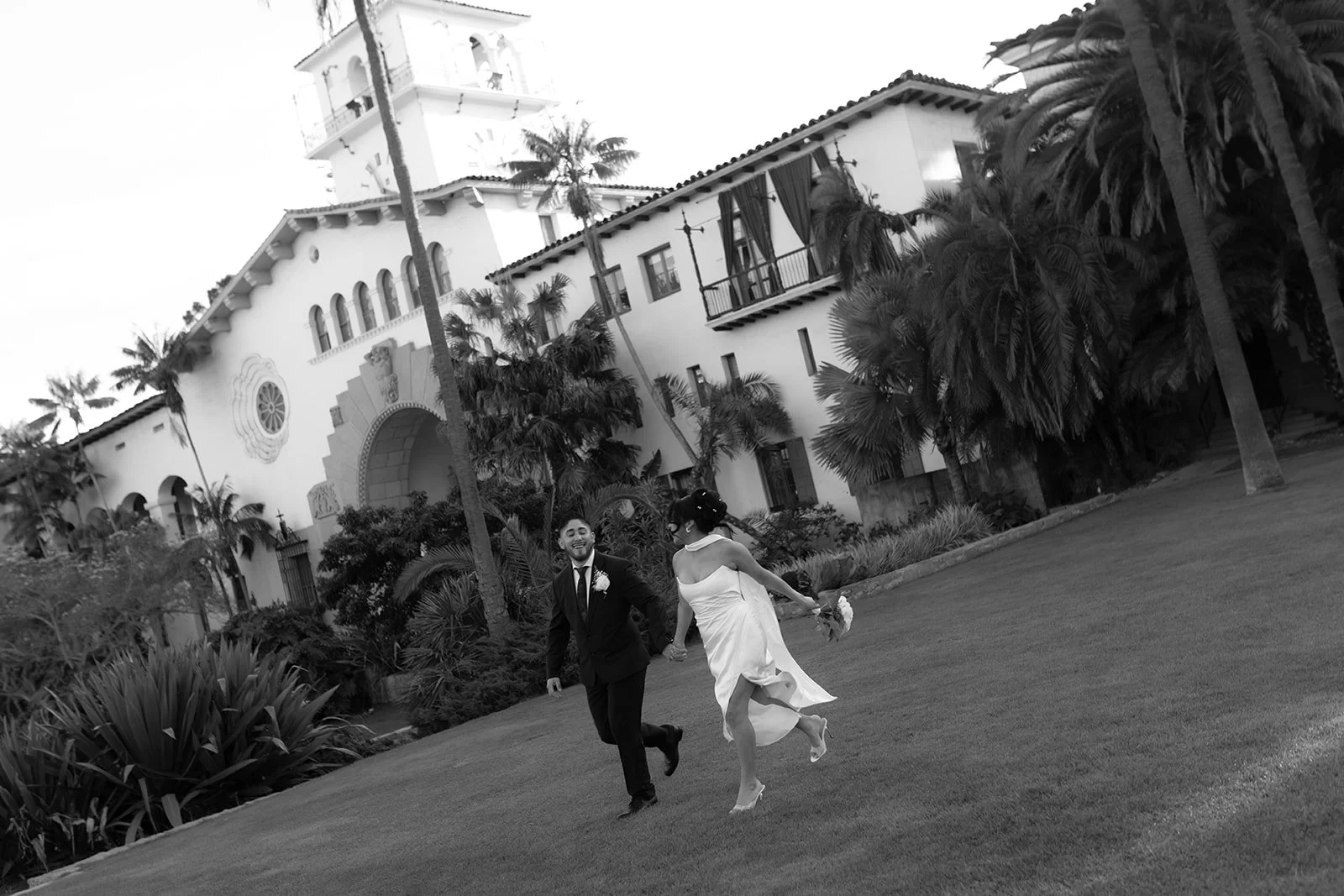 Black and white candid of couple running across the grass in the gardens at the Santa Barbara Courthouse Sunken Gardens, smiling at each other in a joyful candid moment