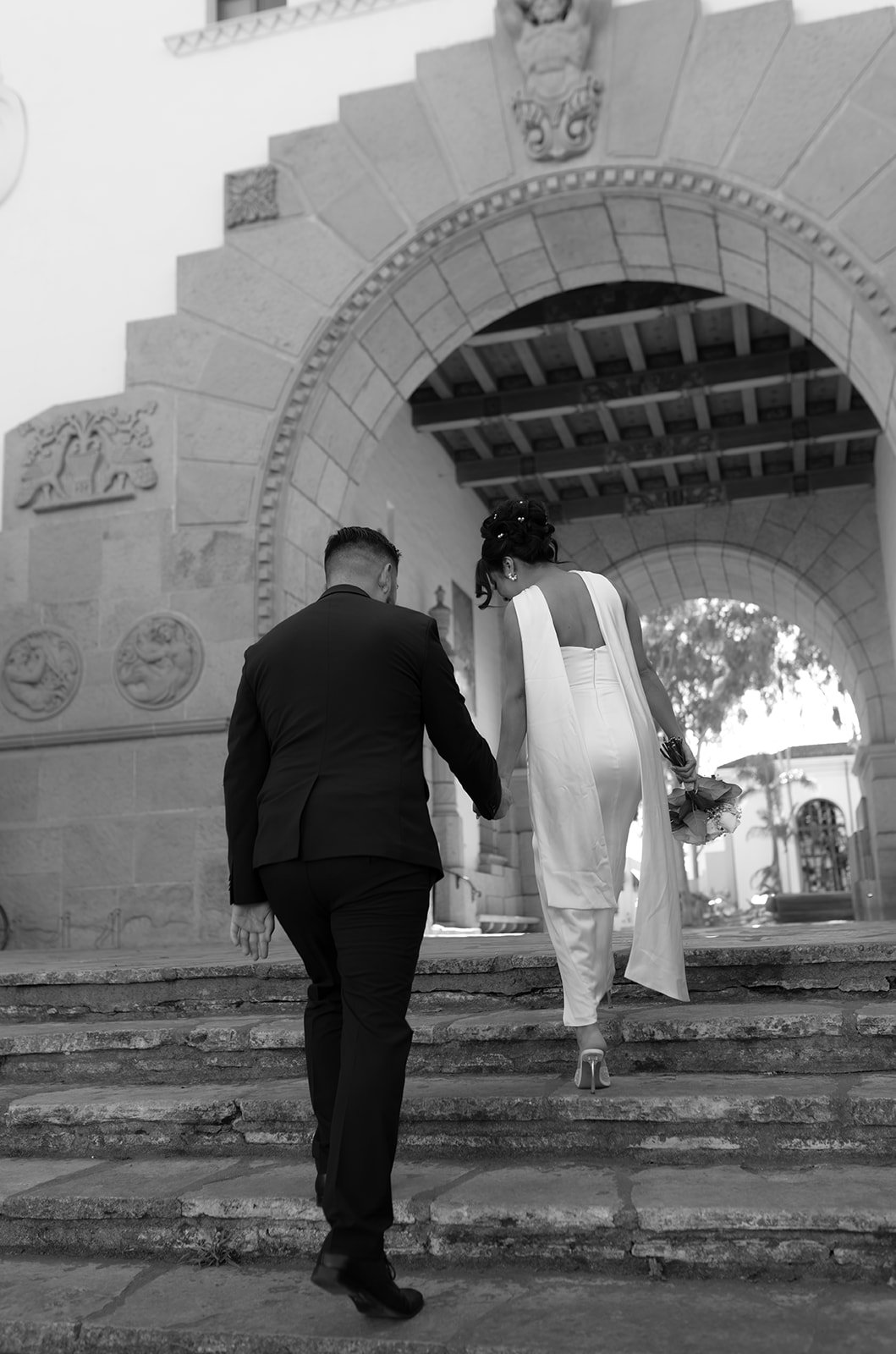 Black and white candid photo of a couple holding hands while walking down the Santa Barbara Courthouse stone stairs, captured mid-step with natural movement and soft, documentary-style intimacy.
