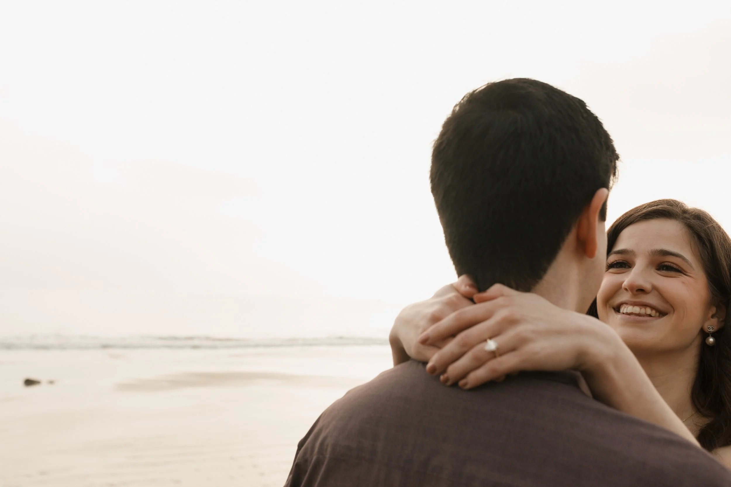 close up photo of couple showing off ring from proposal at newport beach. guys head is towards camera while girl has arms wrapped around his neck smiling at him