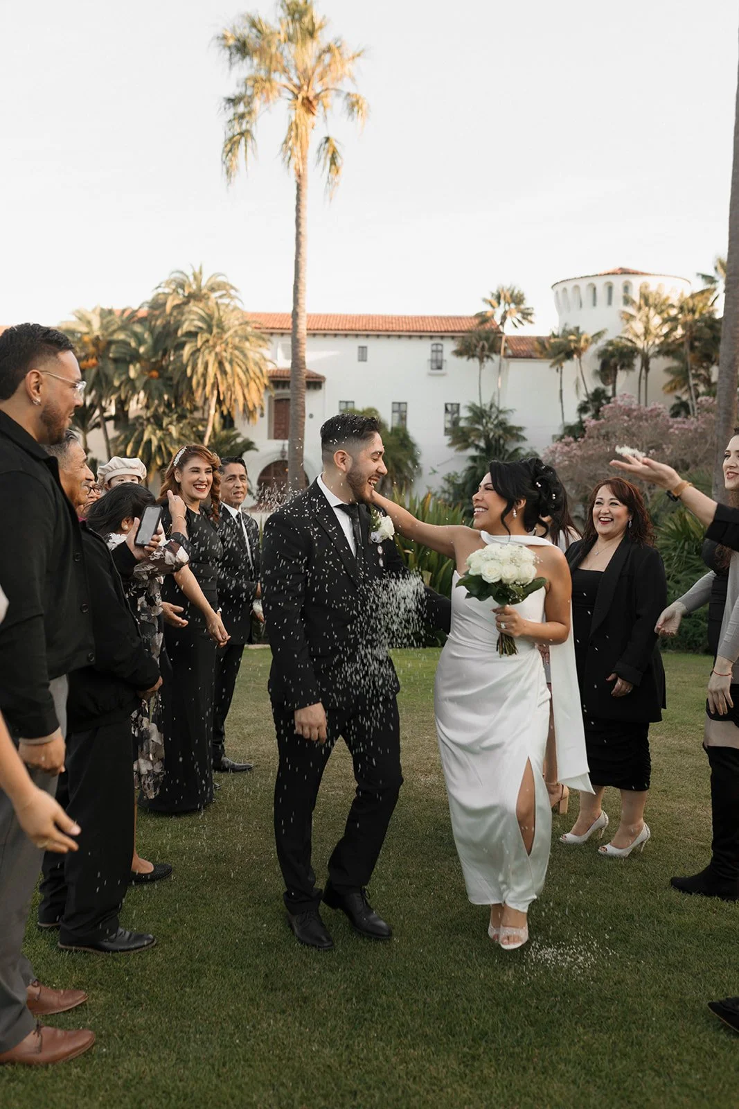 Candid moment of couple smiling and holding each other as they walk through a tunnel of family at the Santa Barbara Courthouse Sunken Gardens, with guests throwing rice in celebration in the background