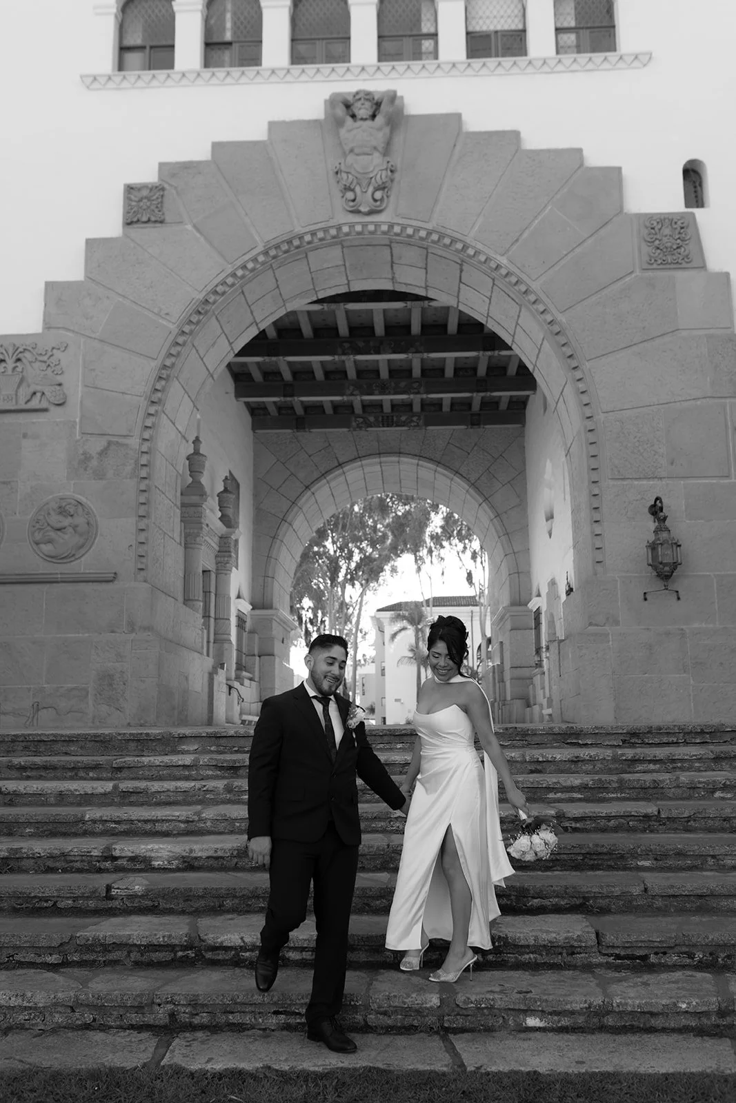 Black and white candid photo of a couple holding hands while walking down stone stairs, captured mid-step with natural movement and soft, documentary-style intimacy.