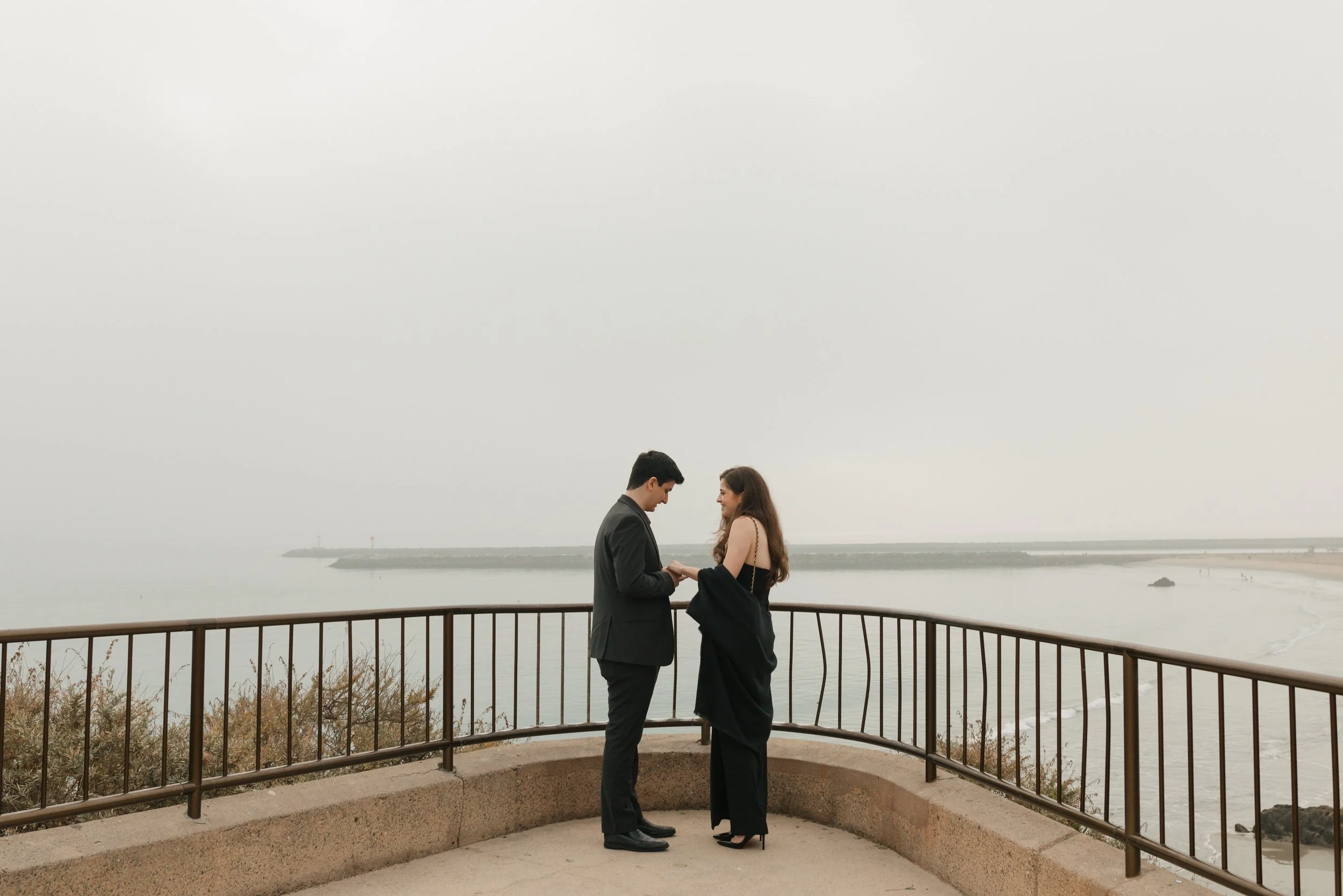 couple at the look out in newport beach for proposal facing eachother with beach in background