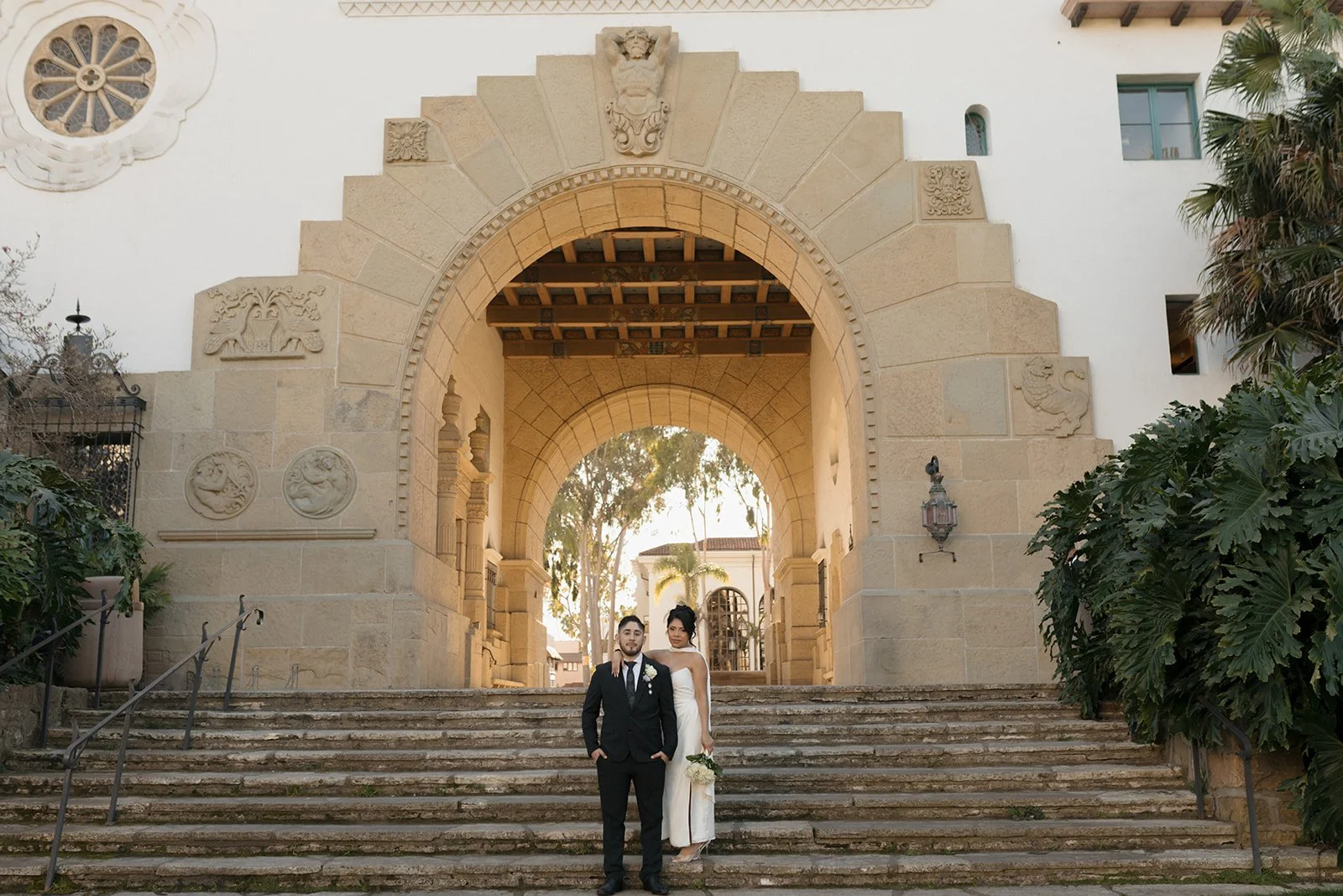 Editorial-style photo of a couple posed on the stairs in front of the Anacapa Arch, capturing a romantic, cinematic elopement moment with intentional posing, elegant composition, and the iconic coastal architecture framing them.