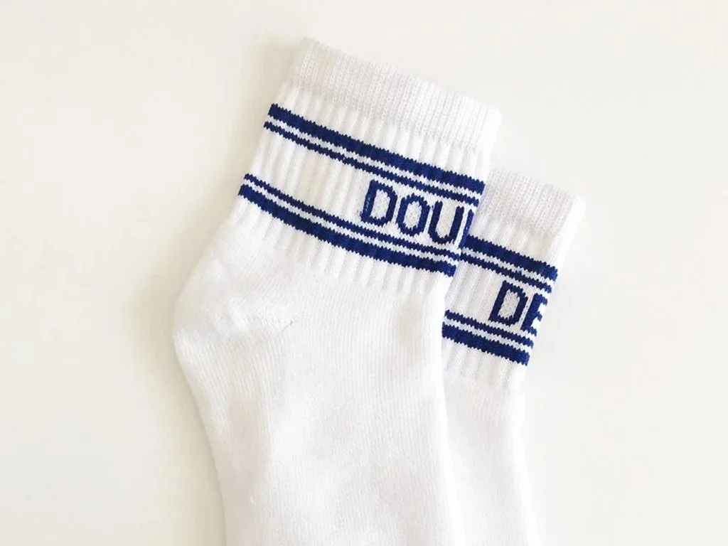 A pair of retro white athletic socks with blue horizontal stripes and the words DOUBLE DEUCE in blue on the top part of each sock, laid on a white surface.