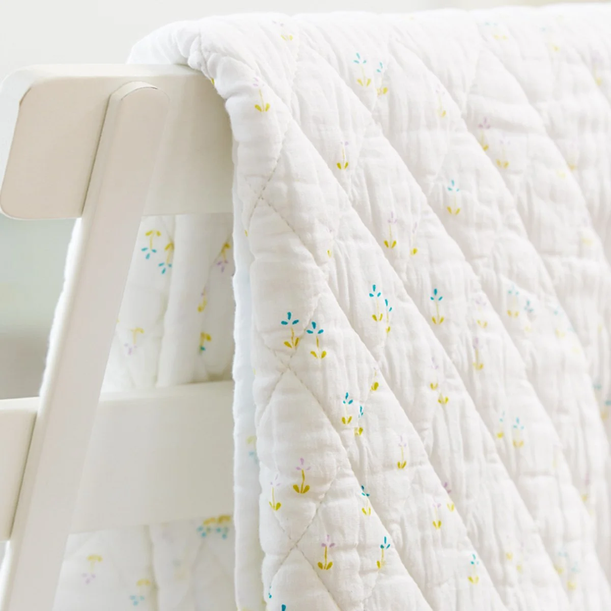 Close-up of a white quilt with small, colorful floral patterns hanging over the side of a white wooden bed frame.