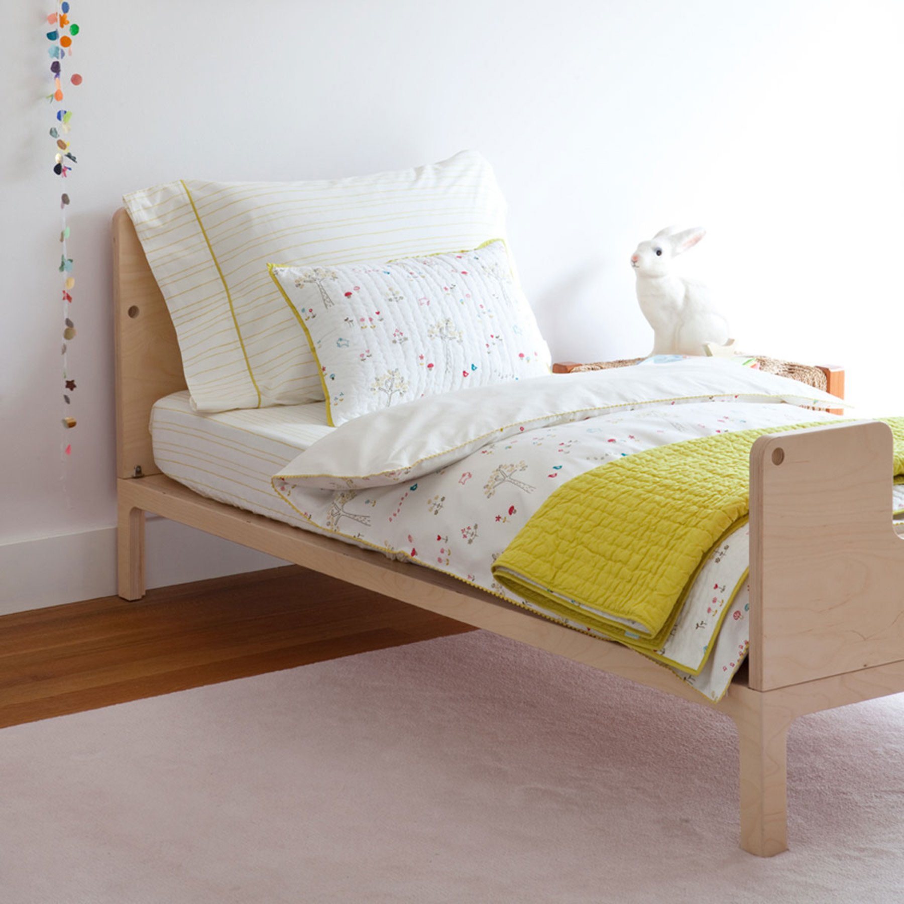A neatly made toddler bed with light wood frame, white and yellow striped pillow, and a white quilt with colorful embroidery, topped with a folded yellow quilt. Part of a cream-colored carpeted floor is visible.