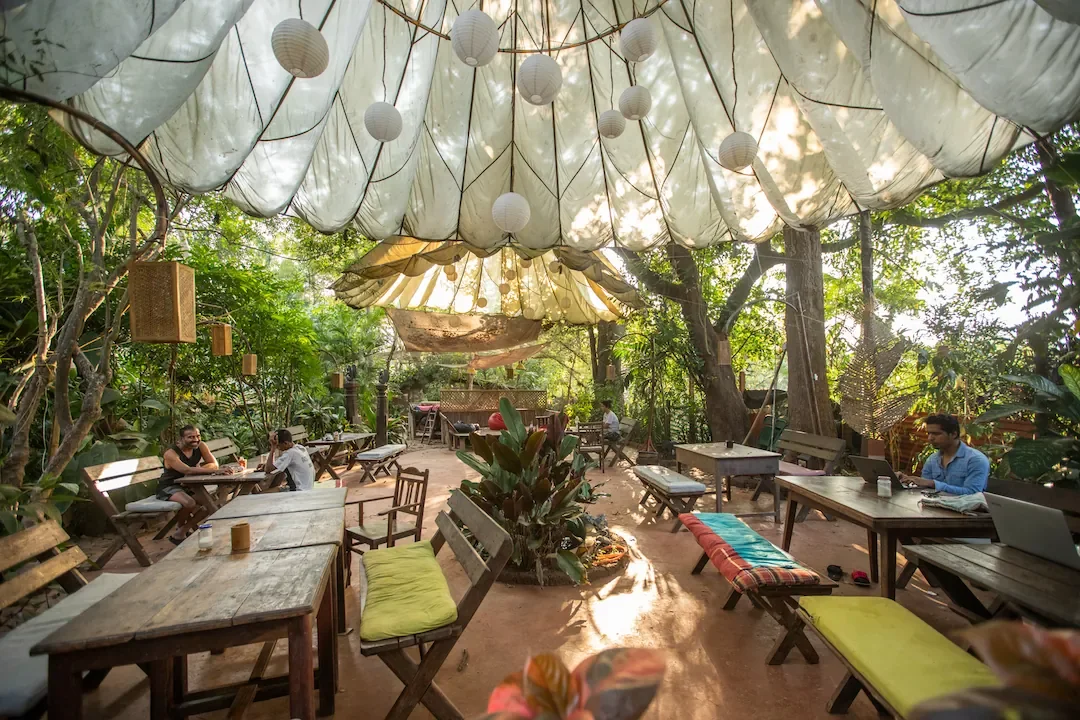 Charming outdoor café with wooden tables and chairs, hanging paper lanterns and fabric drapes, surrounded by lush greenery, inviting relaxed dining in nature.
