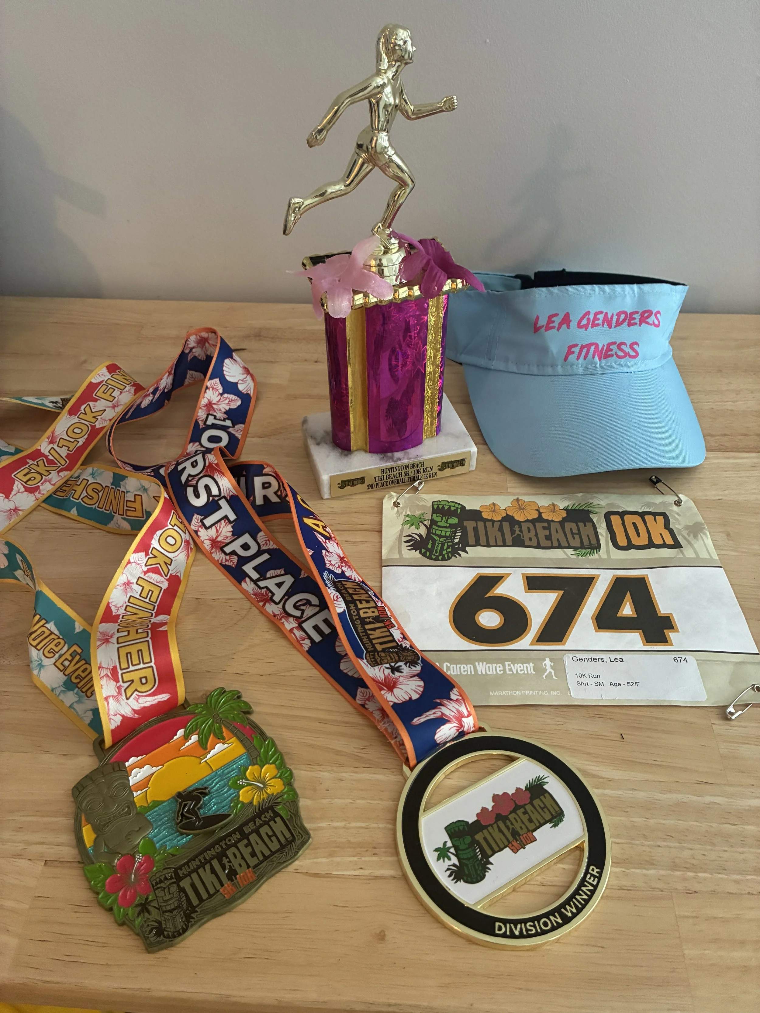  Tiki Beach 10K finisher medal, Division winner medal, and overall female 2nd place trophy along with racer’s bib and Lea Genders fitness visor. 
