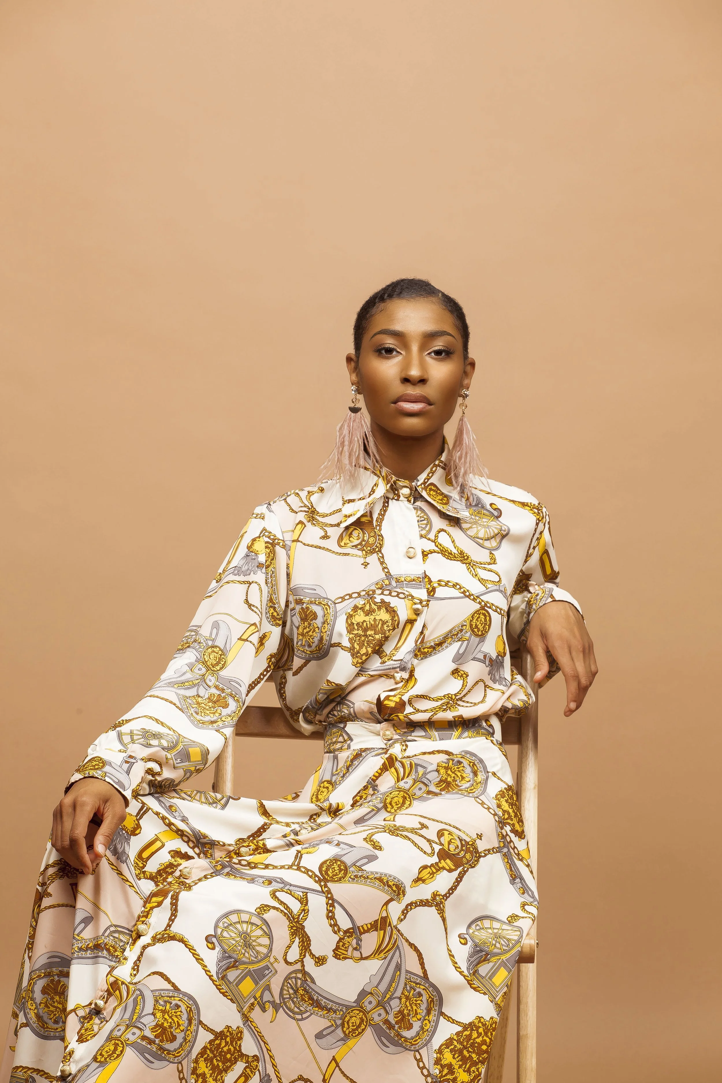 A woman with dark skin and short black hair, wearing a white and gold patterned silk shirt and matching skirt, sitting on a wooden chair against a beige background.