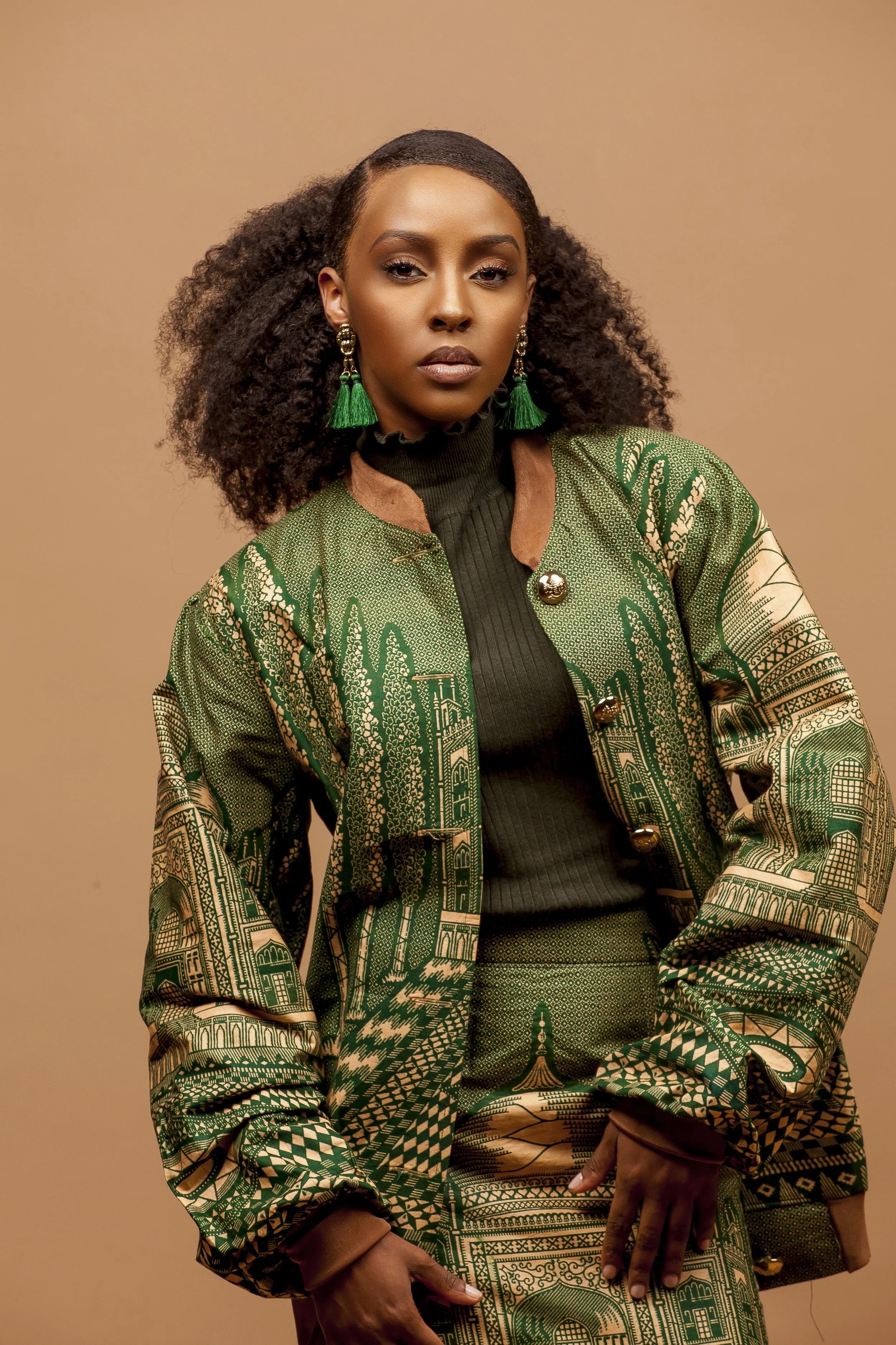 Portrait of a woman with dark skin and curly hair wearing large green tassel earrings, a green patterned jacket with gold buttons, and matching patterned skirt, set against a beige background.