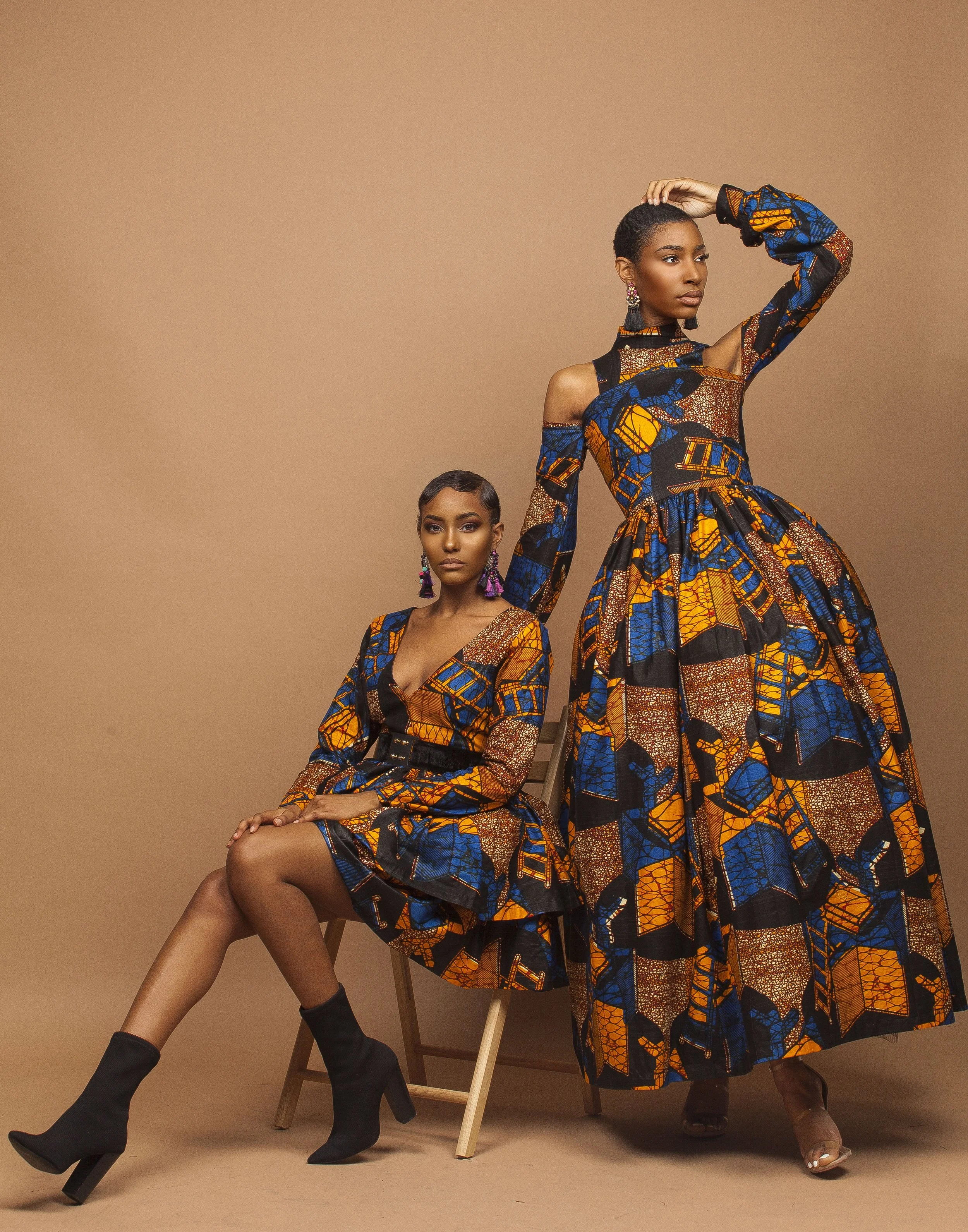 Two women in vibrant African print dresses pose against a beige background. One seated with crossed legs, the other standing with one arm raised, both wearing earrings and black boots.