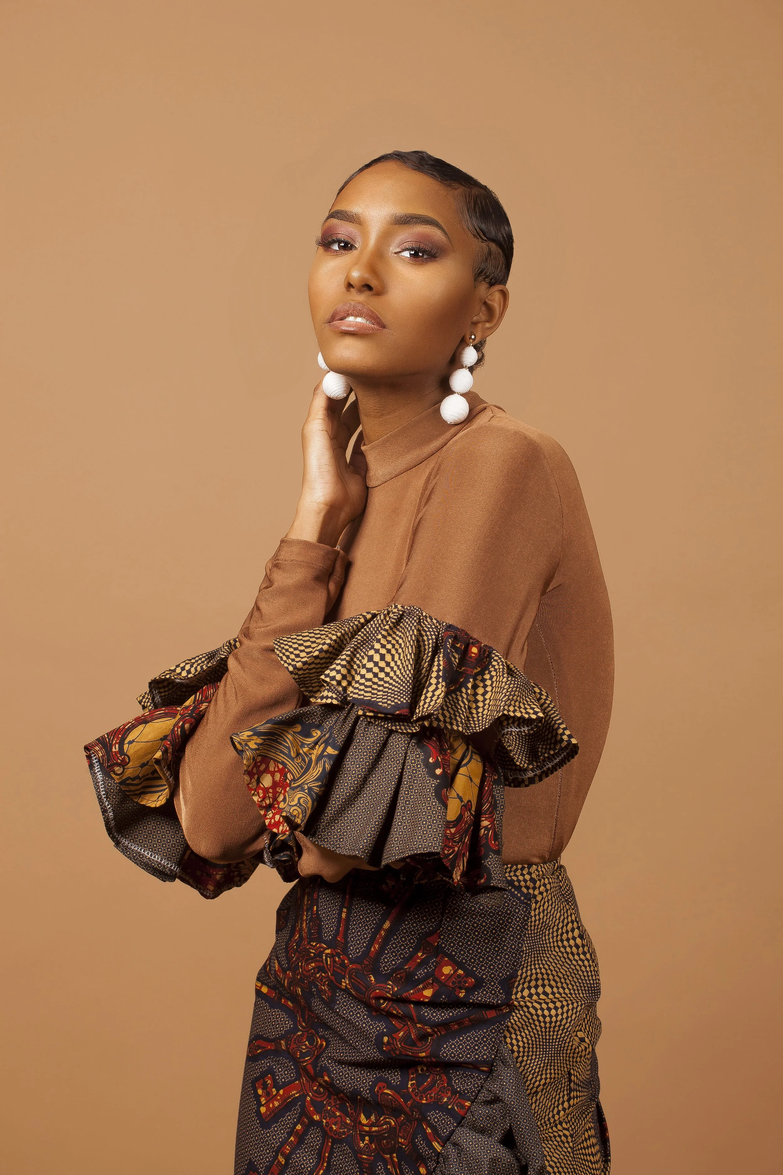 Portrait of a woman wearing large white earrings, a brown top, and a patterned fabric accessory. She has short dark hair, glowing makeup, and a confident expression against a beige background.