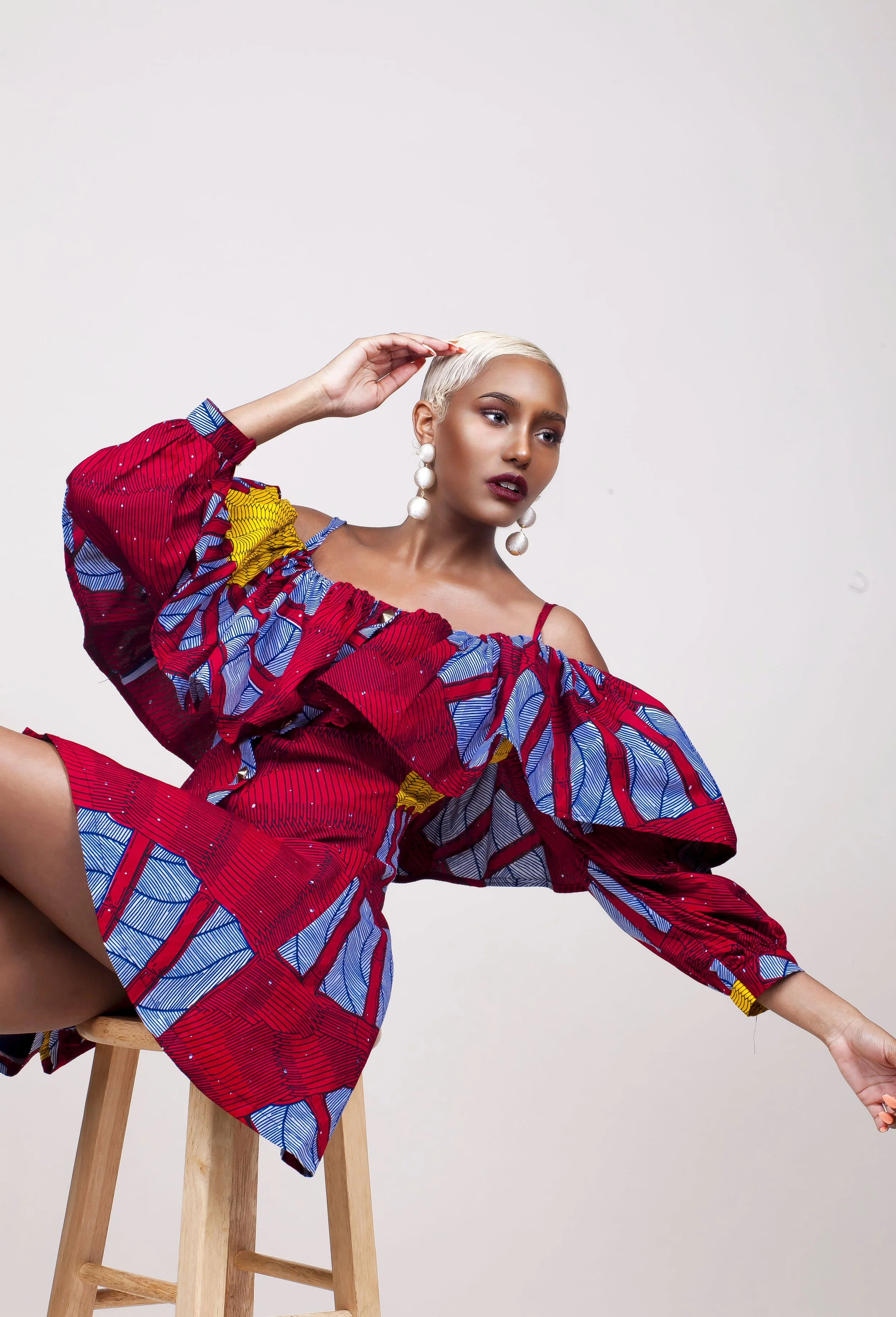 A woman with short platinum blonde hair, wearing large pearl earrings, a red, blue, yellow, and black patterned off-shoulder dress, seated on a wooden stool against a plain background.