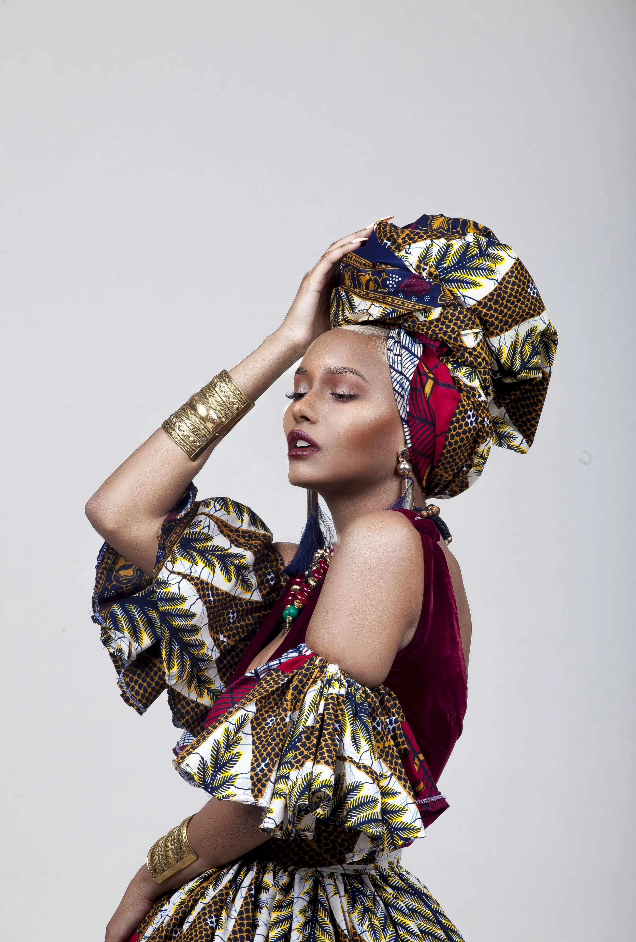 Woman wearing African print fabric and jewelry, with a headwrap, posed against a plain background.
