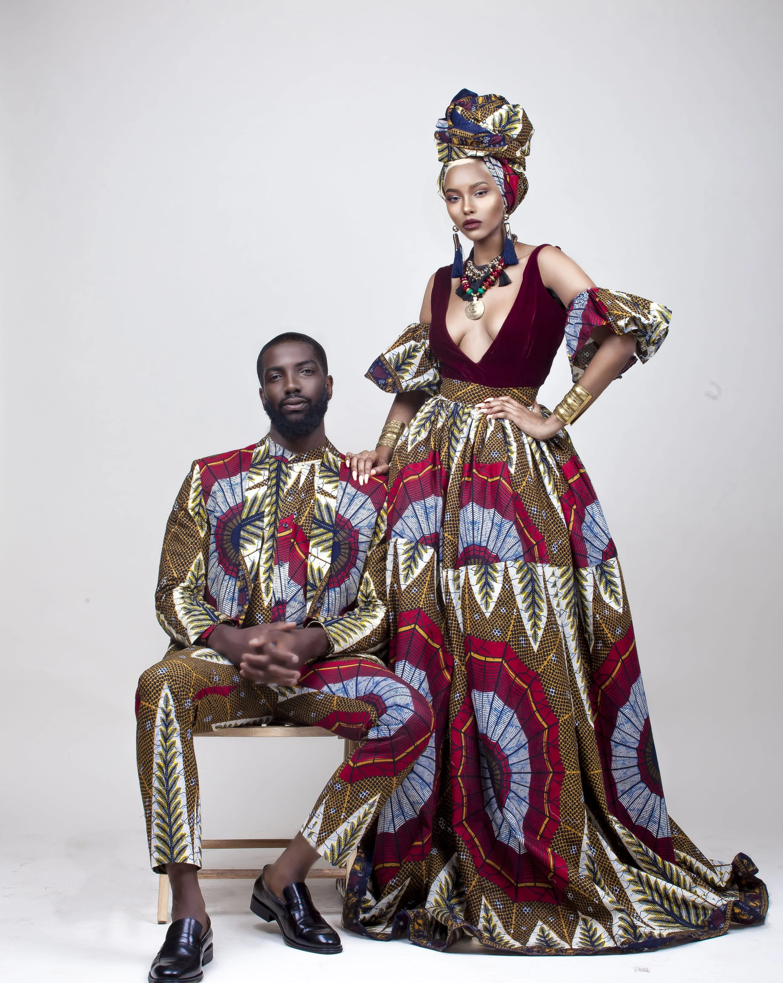 A woman and a man dressed in traditional African attire with vibrant patterns. The woman wears a large headwrap, jewelry, and a flowing dress, while the man wears a matching shirt and pants, seated on a wooden stool, against a white background.