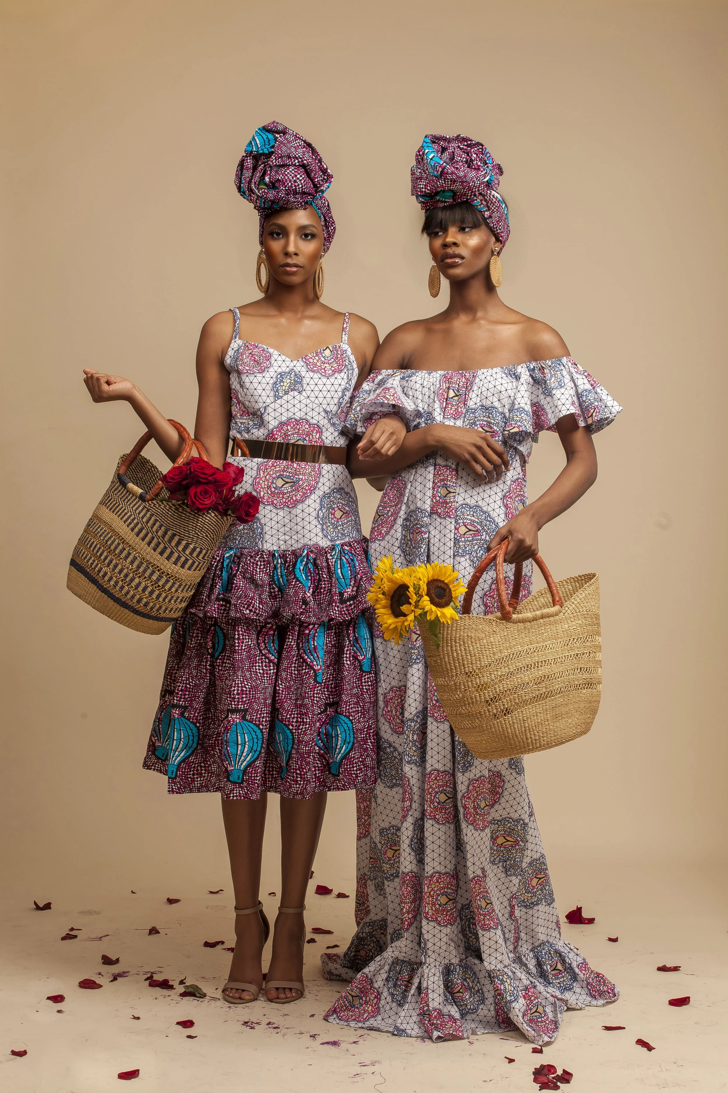 Two women with dark skin wearing matching colorful African print dresses and head wraps pose against a beige background. They hold woven straw bags, one with red roses and the other with sunflowers. The floor is scattered with red and brown petals.