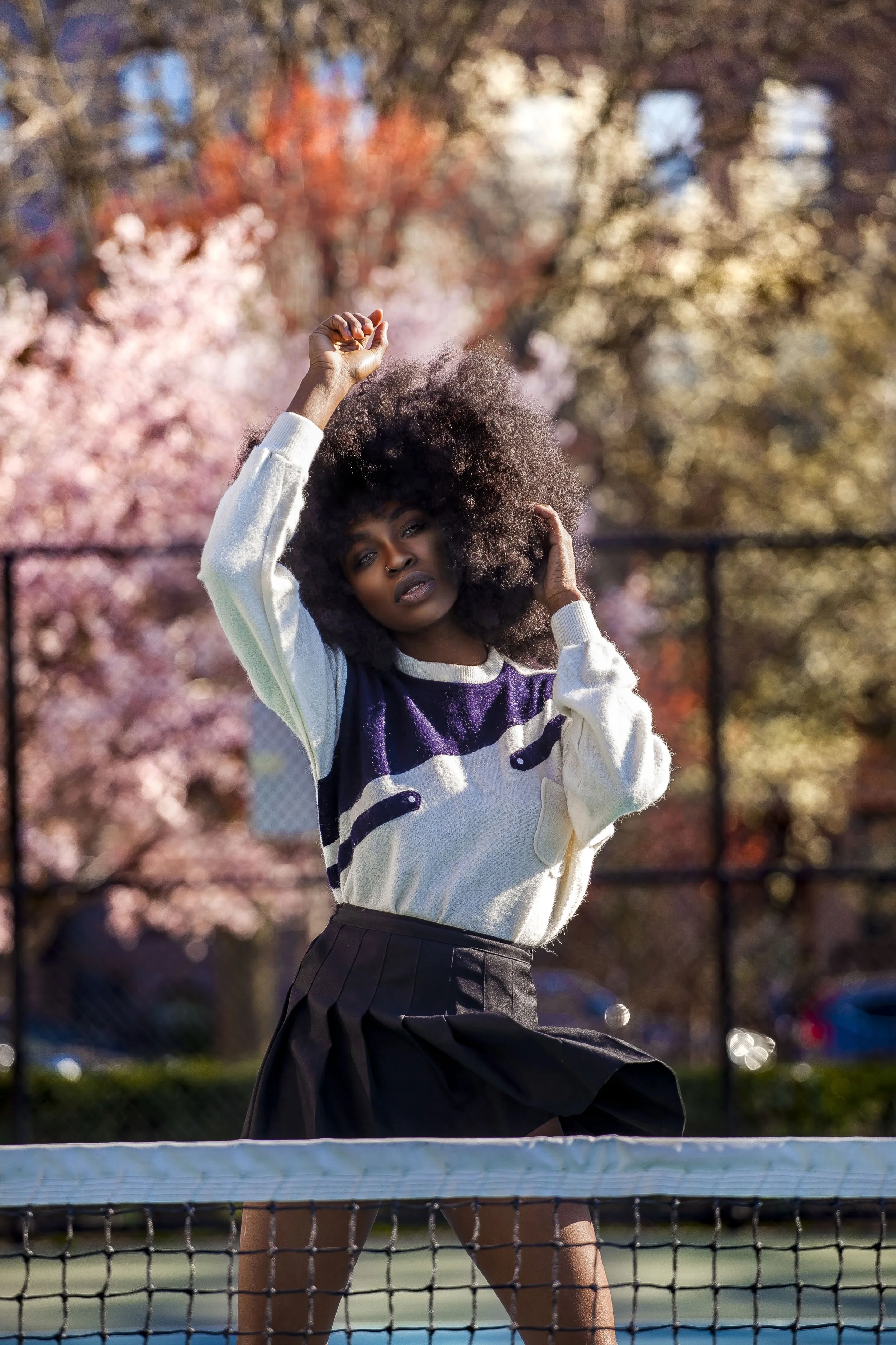 A woman with voluminous curly hair wearing a patterned sweater and a black skirt playing tennis on an outdoor court, with autumn trees in the background.