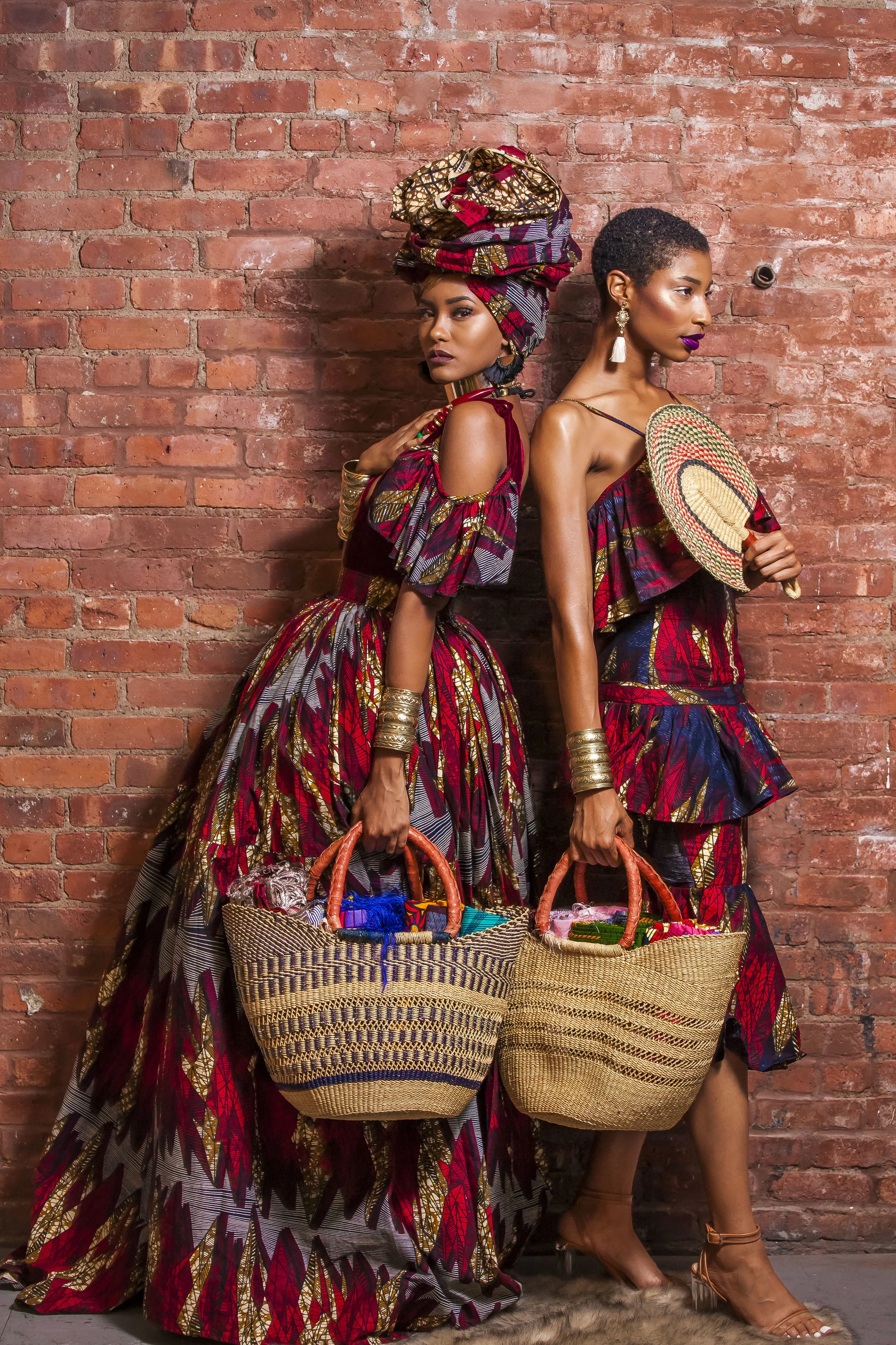 Two women dressed in colorful African-inspired clothing and accessories standing against a brick wall. They hold large woven baskets filled with various items. One woman wears a headwrap and statement earrings, while the other has short hair and hold