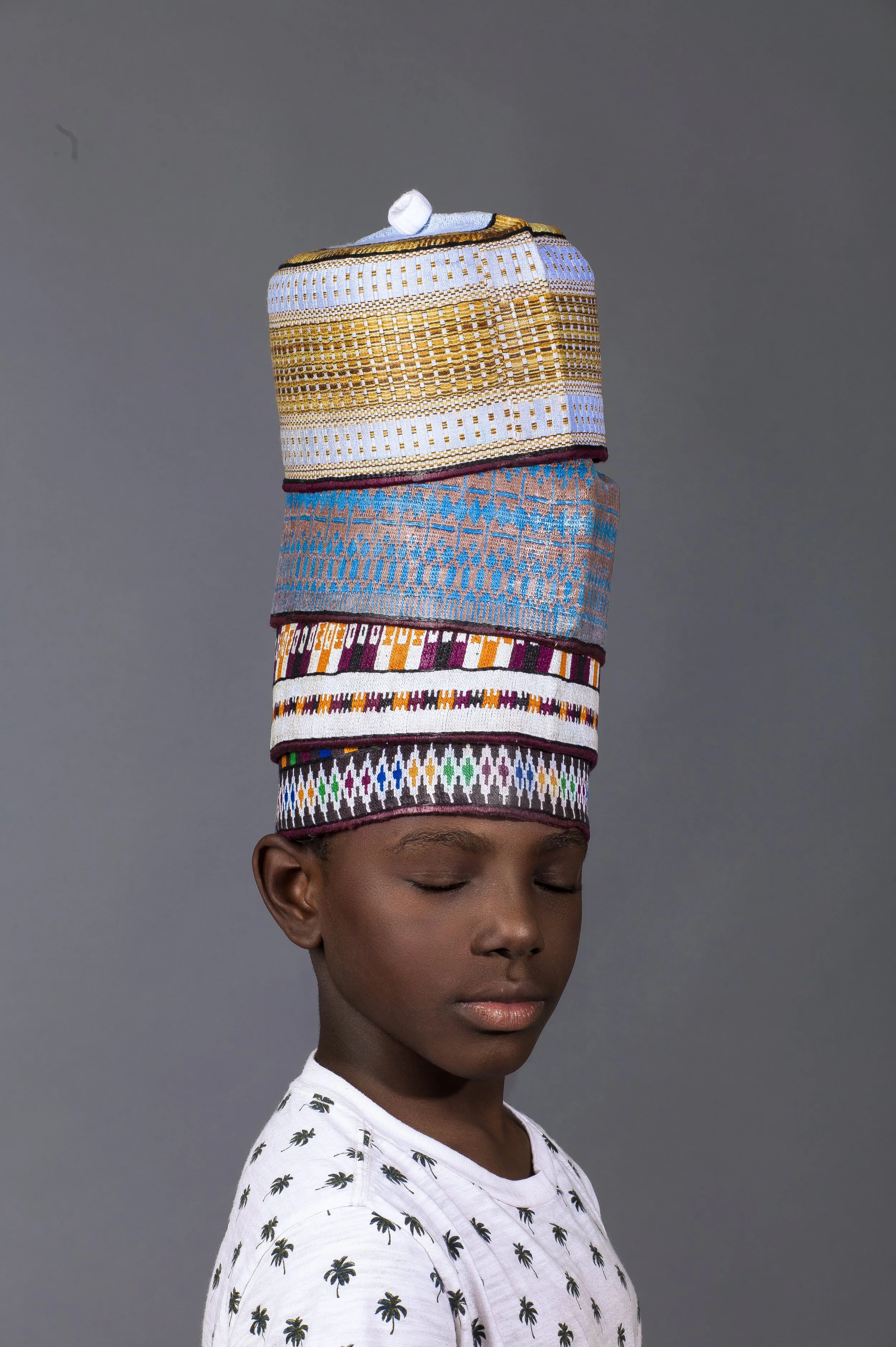 A young person with closed eyes and short hair wearing a white t-shirt with small black palm tree patterns, and a tall, colorful, patterned hat resembling stacked fabric layers in gold, pink, blue, and multicolored designs.
