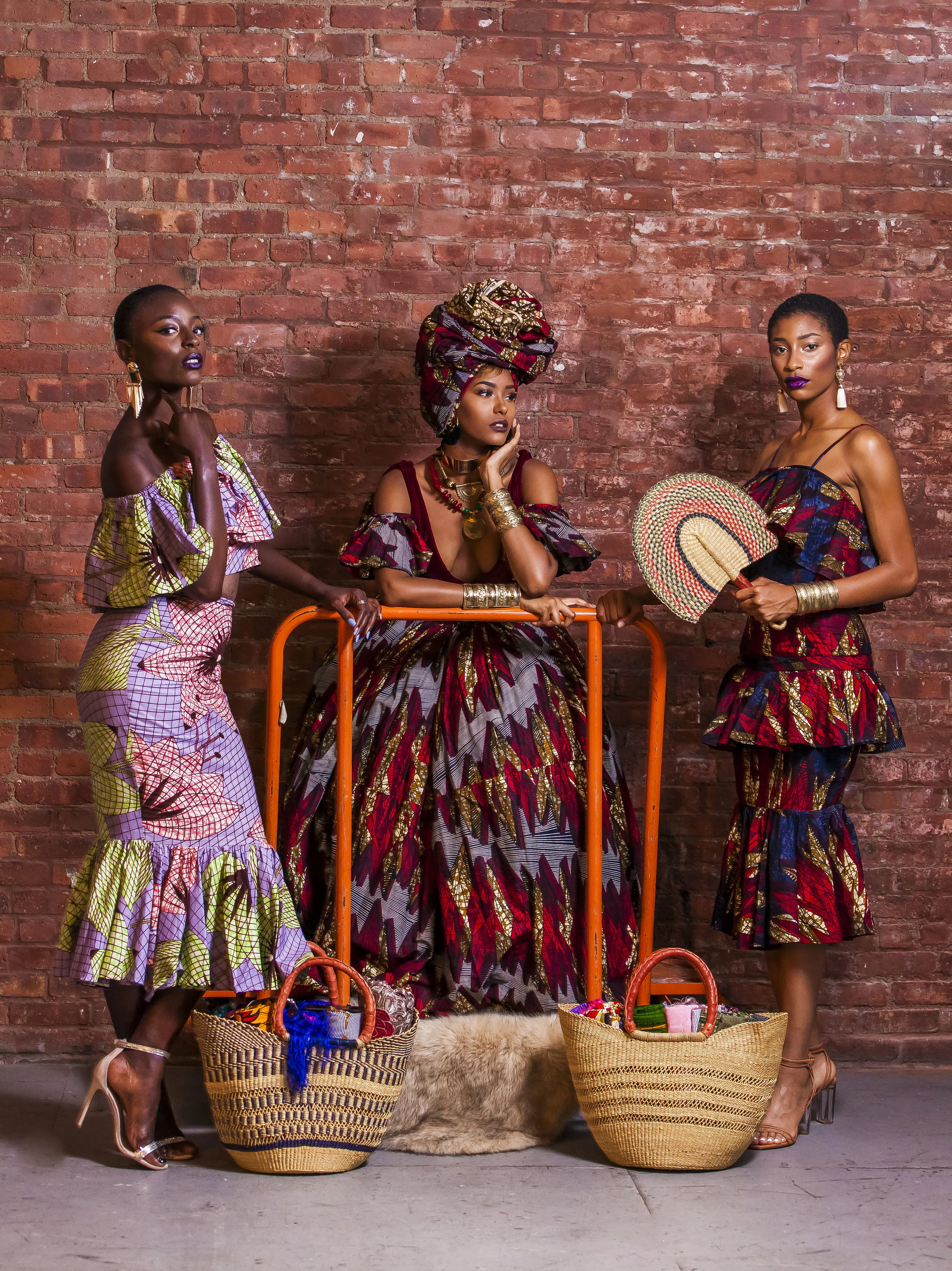 Three women in vibrant, patterned African-inspired dresses and accessories in front of a brick wall. Baskets and handbags are arranged on the floor, and a fuzzy rug is partially visible.