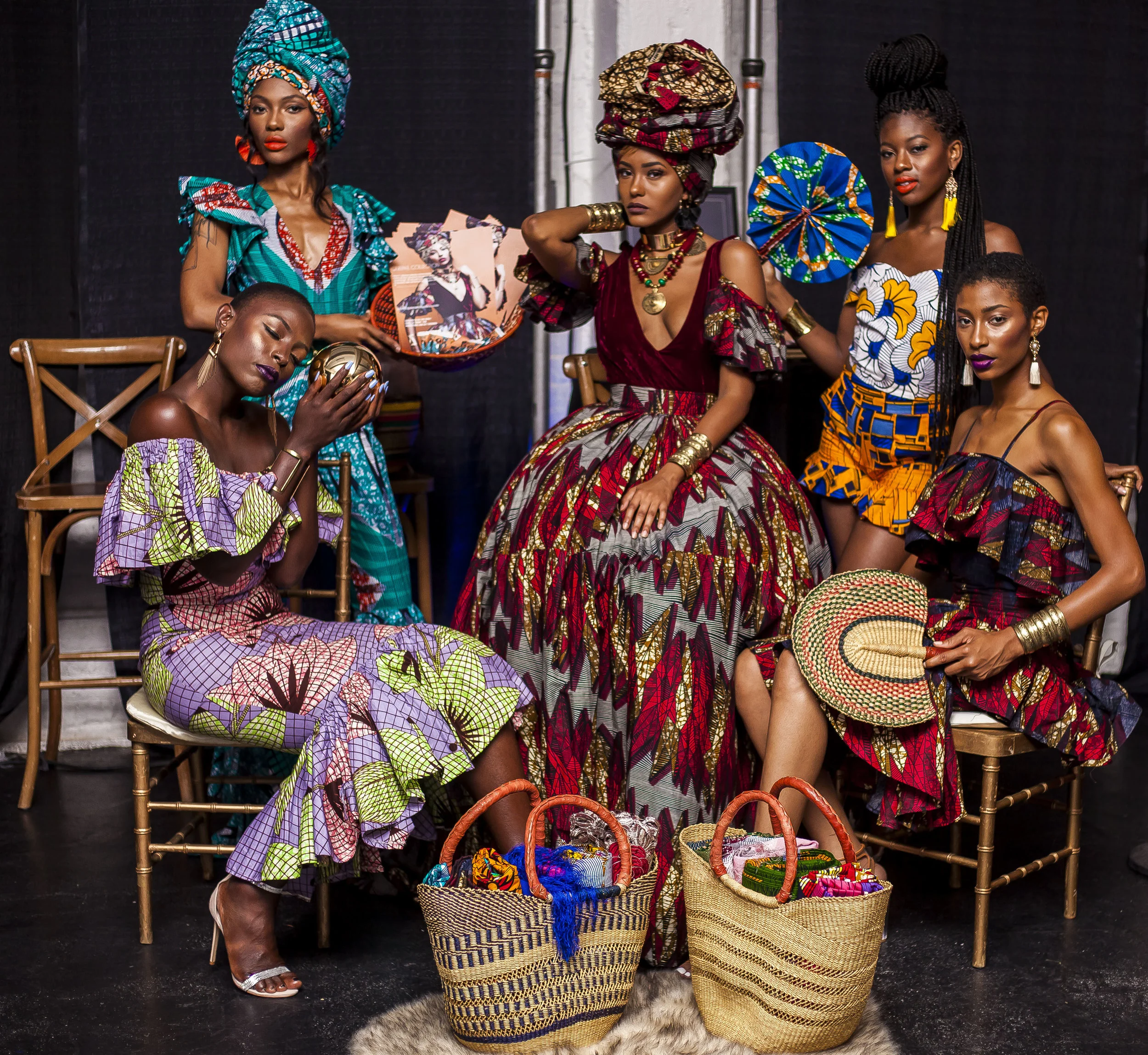 Five women of African descent dressed in colorful African print clothing and traditional accessories, sitting and standing around baskets filled with handmade crafts and textiles, in a room with a black background.
