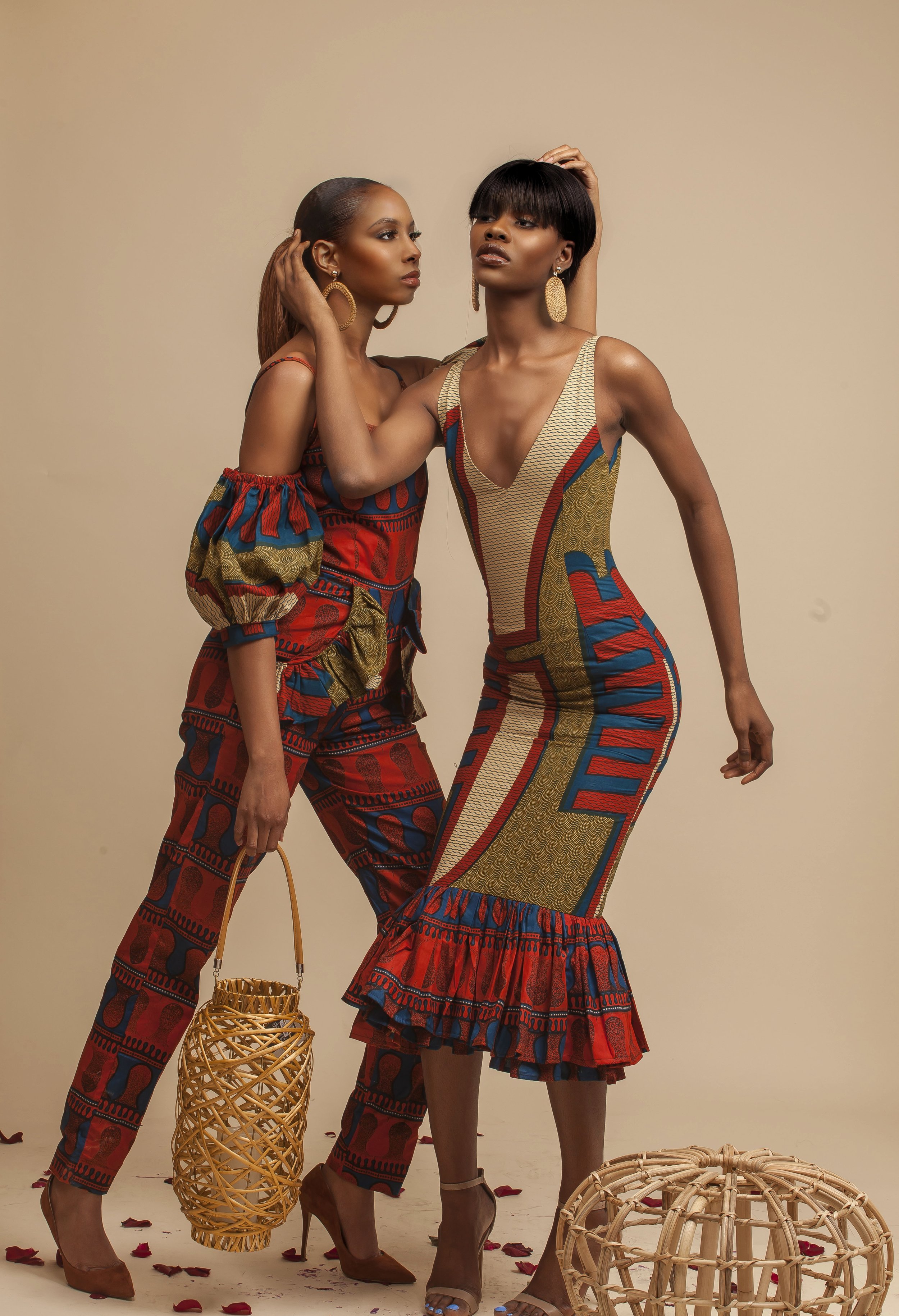 Two women in colorful patterned dresses wearing large earrings, one with her hair in a bun and the other with a short hairstyle, posing against a beige background with wicker baskets on the floor and rose petals scattered around.