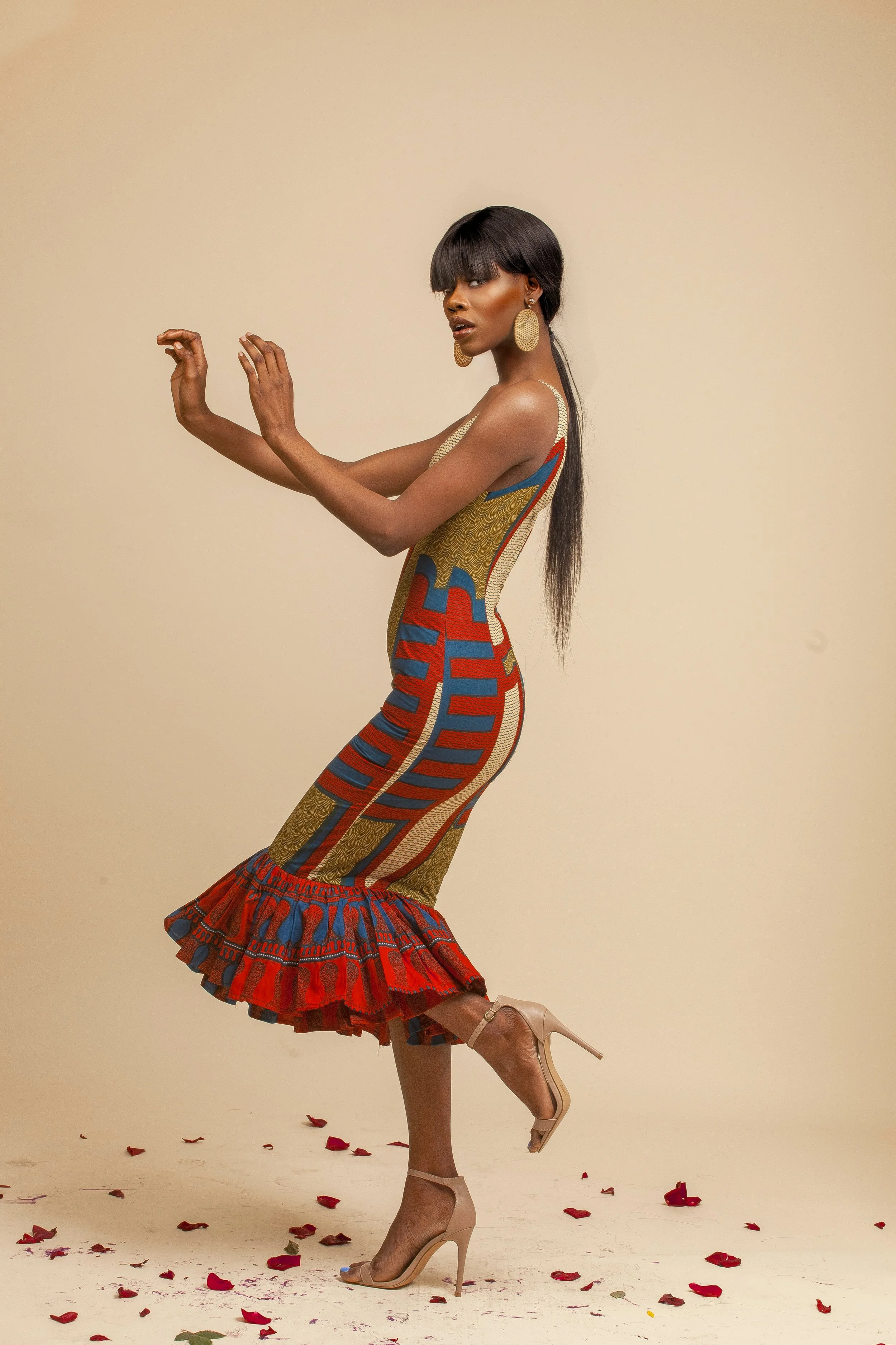 A woman in a colorful patterned dress and beige heels poses on a plain background, surrounded by scattered red and pink flower petals.
