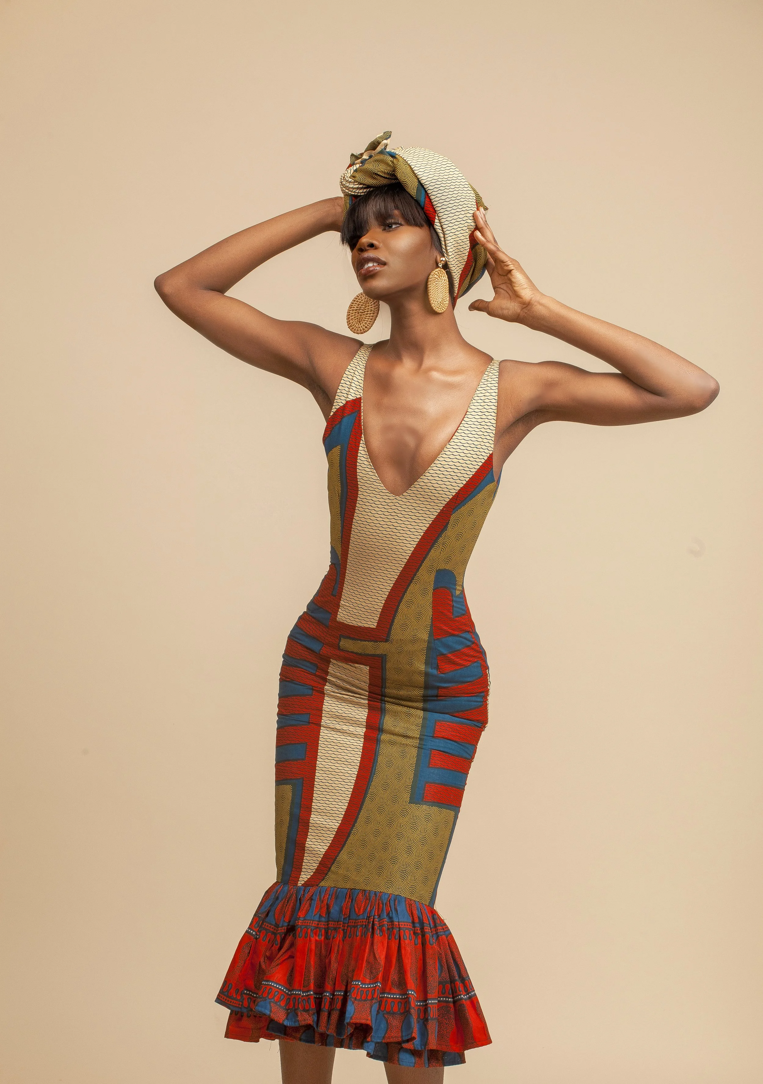 Woman wearing a colorful patterned dress, headwrap, and large earrings, posing with hands in her hair against a beige background.