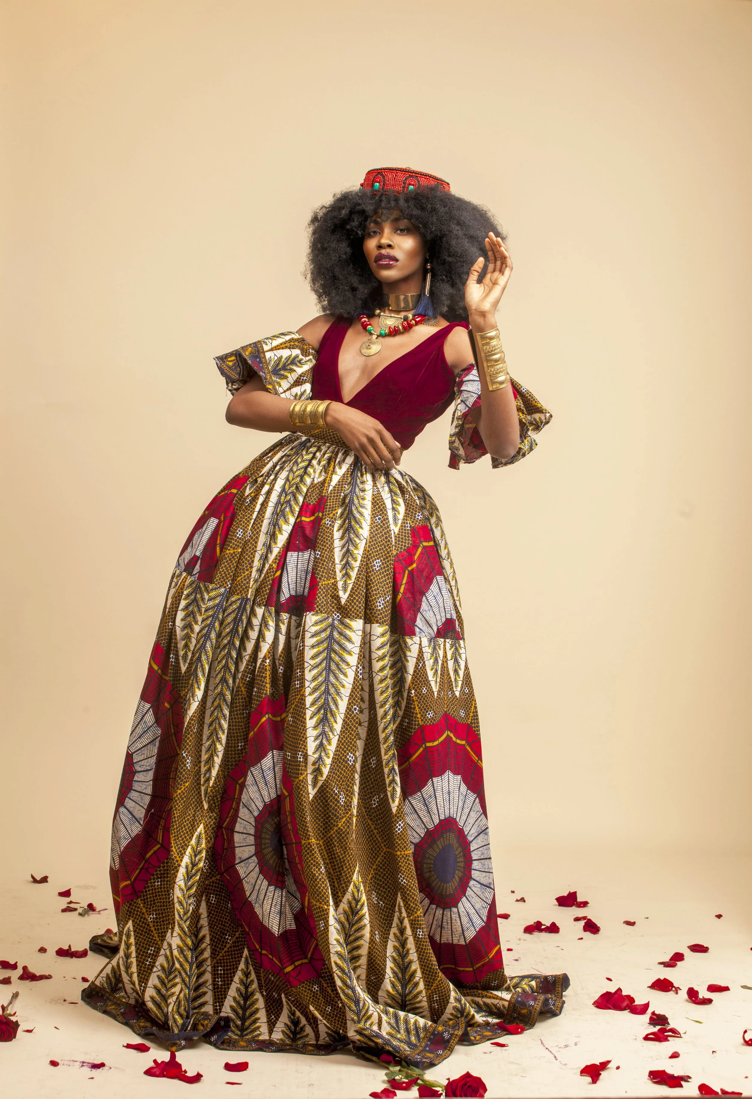 A woman wearing a vibrant, patterned traditional African dress with a deep red top, gold jewelry, and a decorative headwrap stands against a neutral background. She has a confident pose, with one hand raised near her face, and is surrounded by scatte