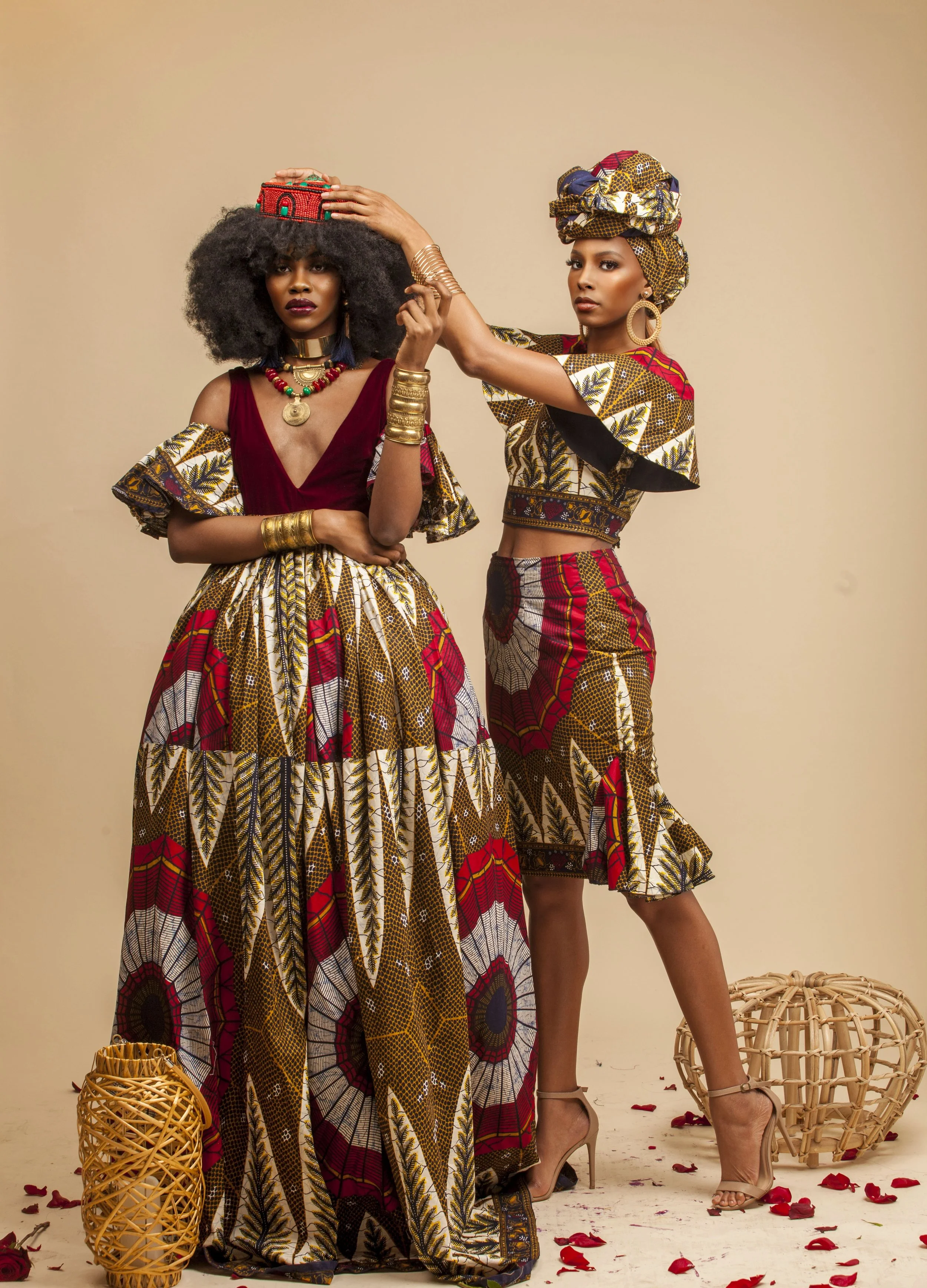 Two women dressed in brightly colored, traditional African-themed clothing standing against a beige background. The woman on the left has voluminous curly hair, wears a red velvet top with orange, red, and gold patterned fabric skirt, and has multipl
