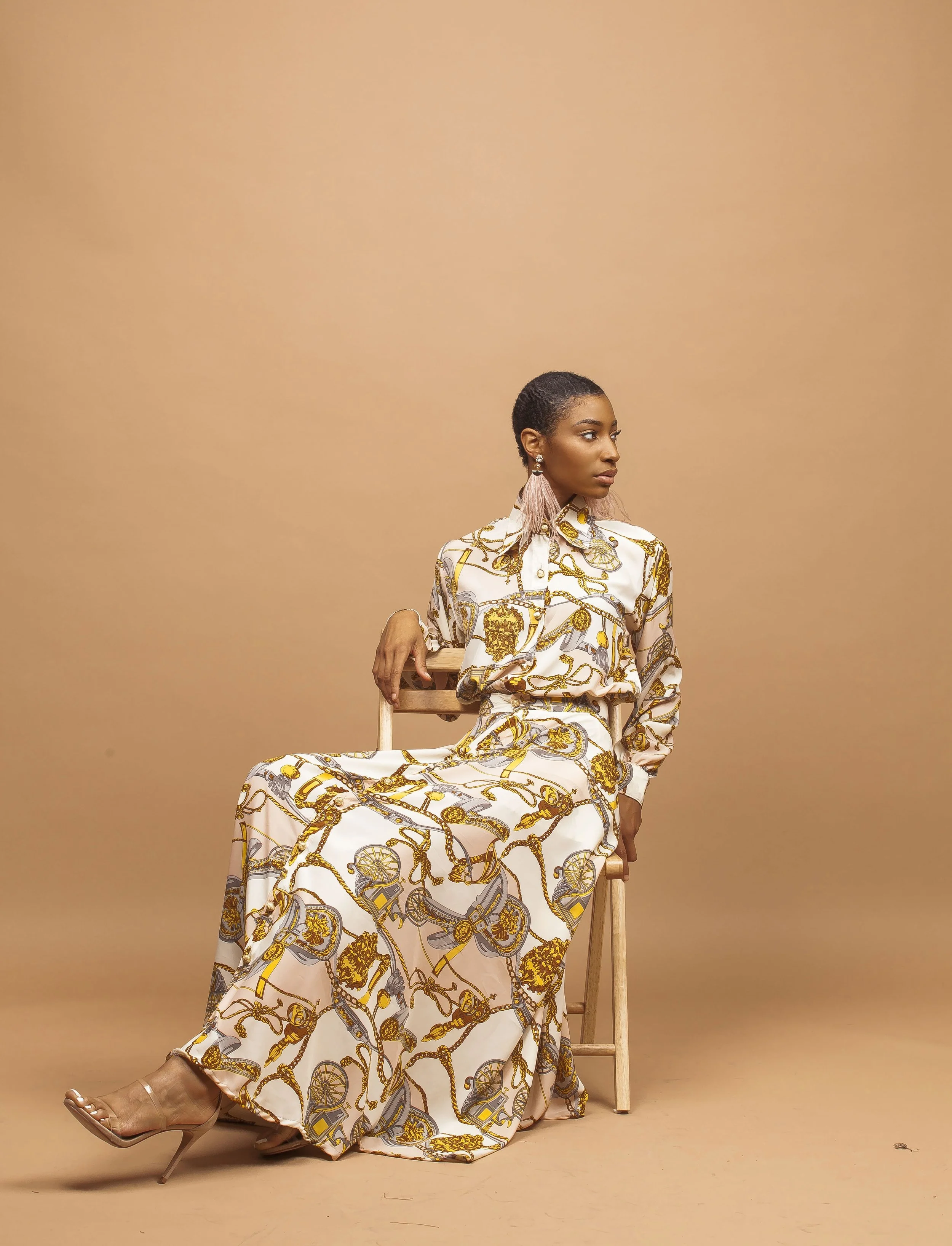 A woman with short hair wearing a gold-printed dress, earrings, and beige high heels sitting on a wooden chair against a beige background.