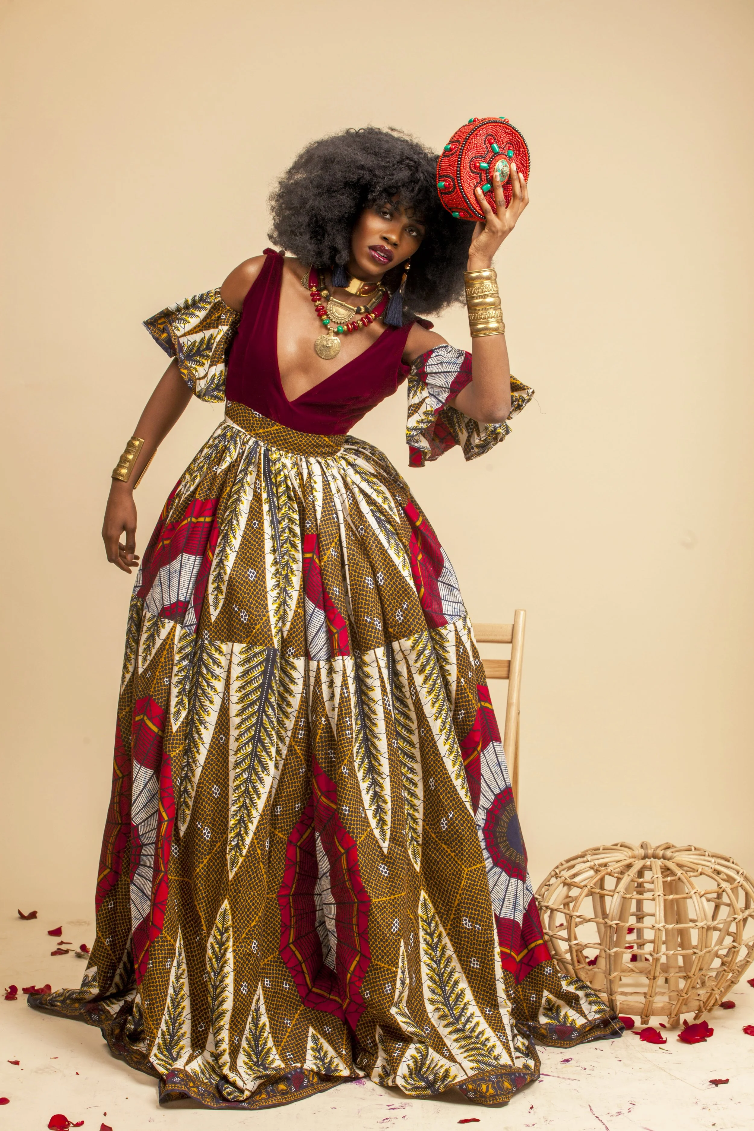A woman with large curly hair wearing a colorful, patterned traditional African dress and jewelry, holding a decorative red clutch, standing in front of a light-colored background with a wicker basket on the floor.