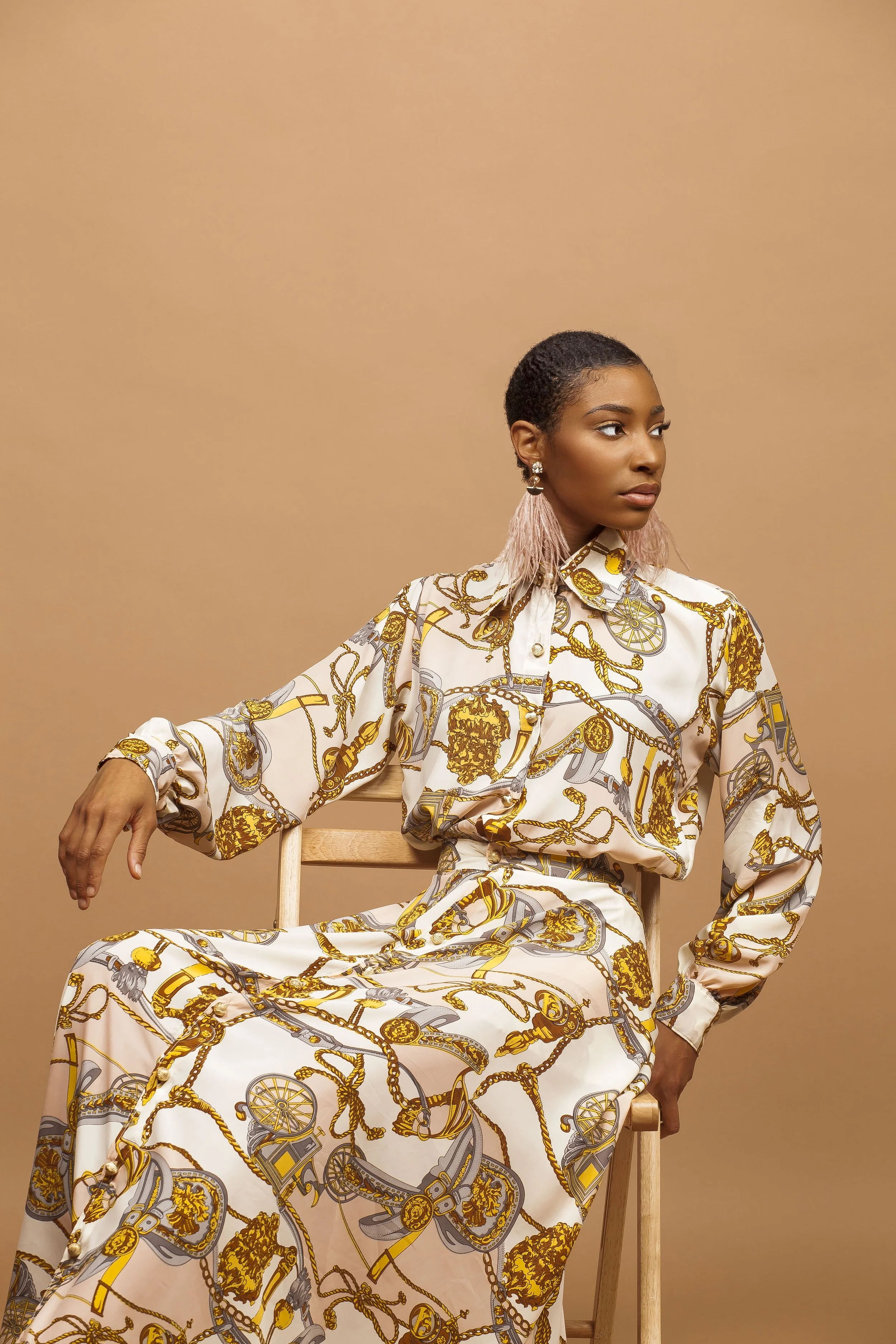 A woman with short dark hair and earrings, wearing a cream and gold patterned dress, sitting on a wooden chair against a plain beige background.