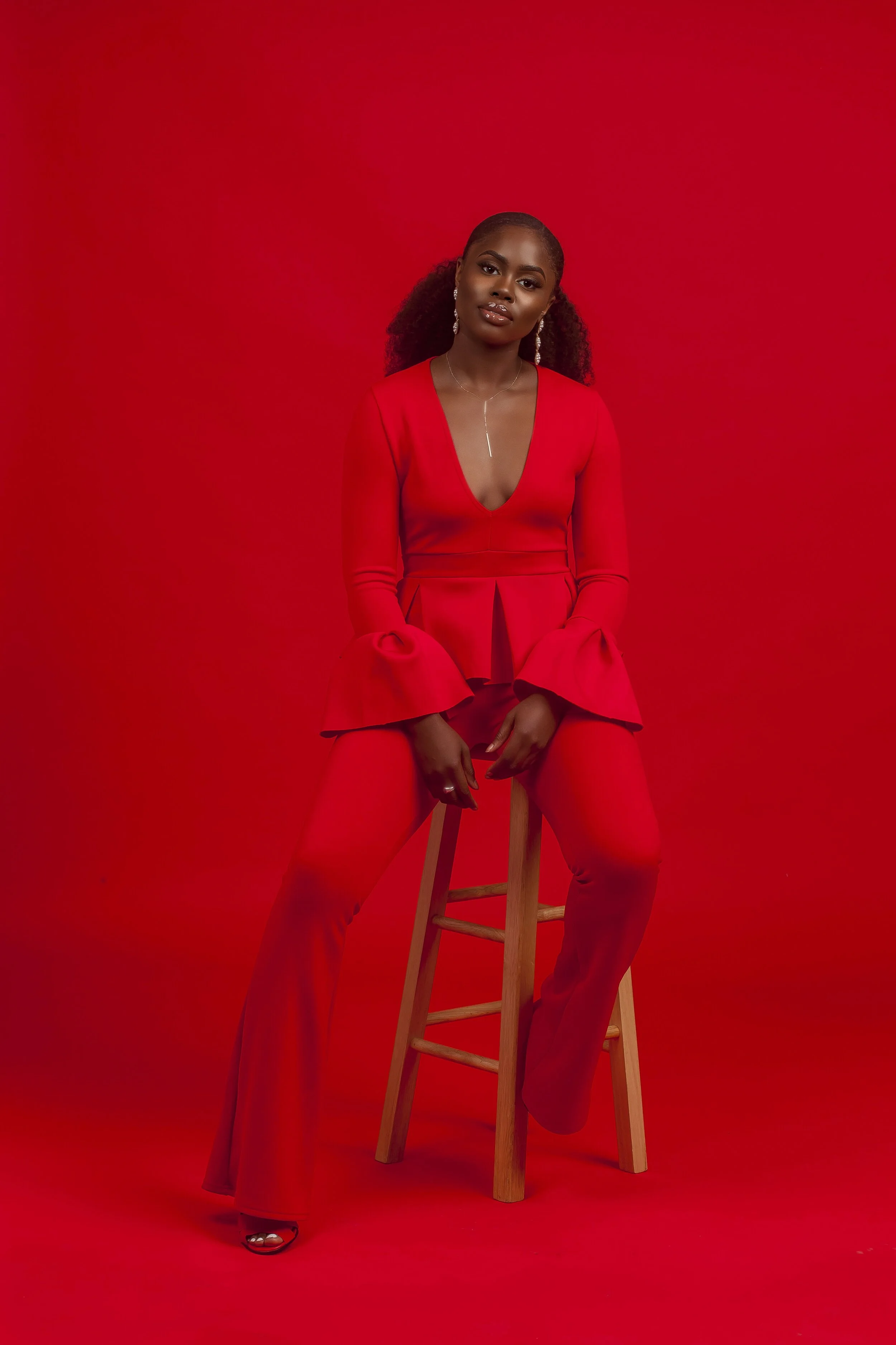 A woman with curly hair tied back, dressed in a red outfit, sitting on a wooden stool against a red background, adorned with jewelry including earrings and a necklace.