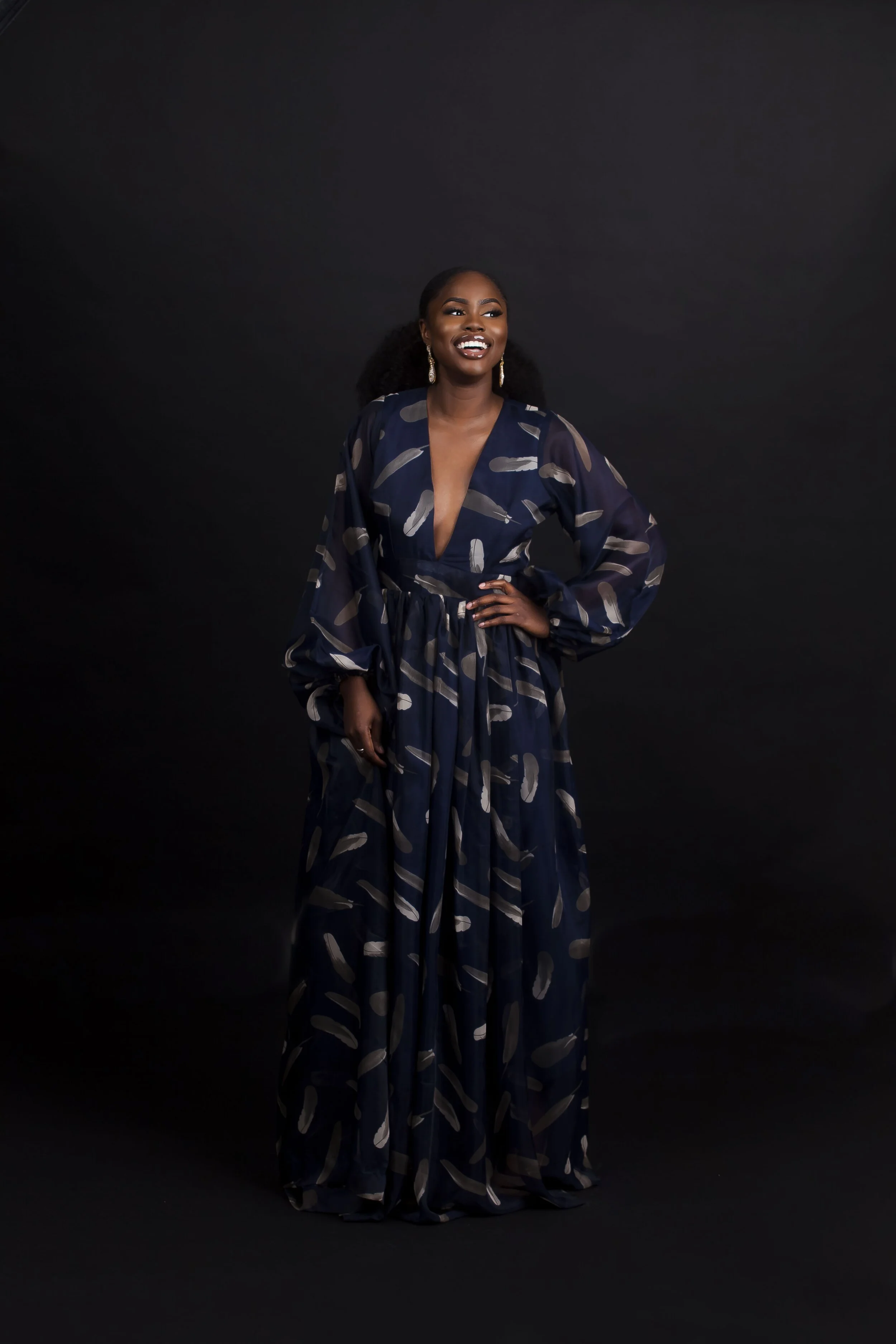 A smiling woman with dark skin, wearing a navy blue dress with a pattern, standing against a black background.