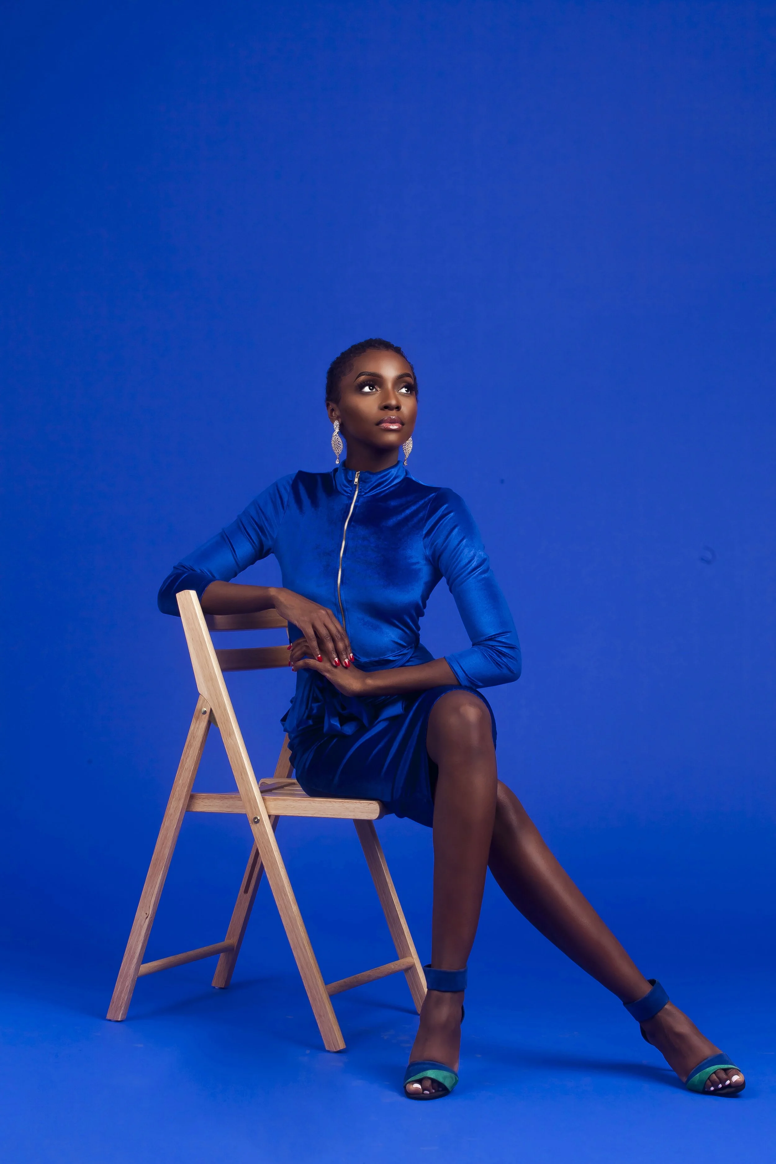 A woman with dark skin in a royal blue dress with a high collar and zipper, sitting on a wooden chair against a matching blue background. She is wearing large, ornate earrings, red nail polish, and turquoise high-heeled sandals. Her hair is pulled ba