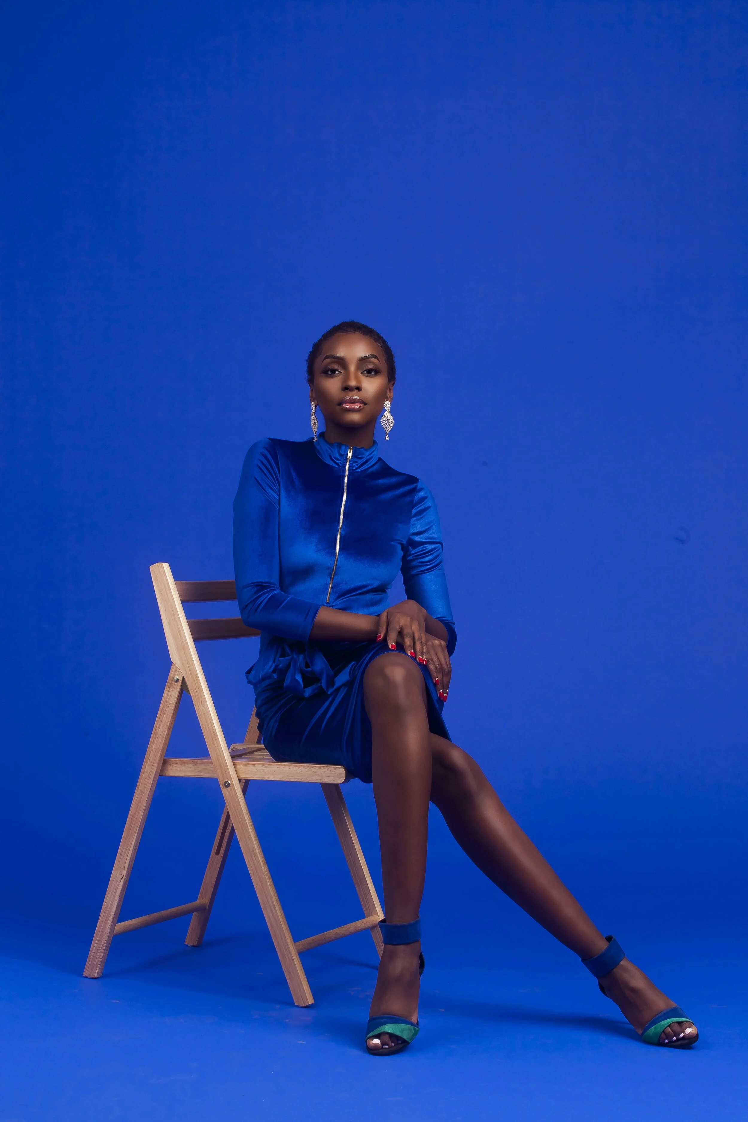 A woman sitting on a wooden chair against a solid blue background, wearing a matching royal blue dress, turquoise high heels, large earrings, and has styled short hair.