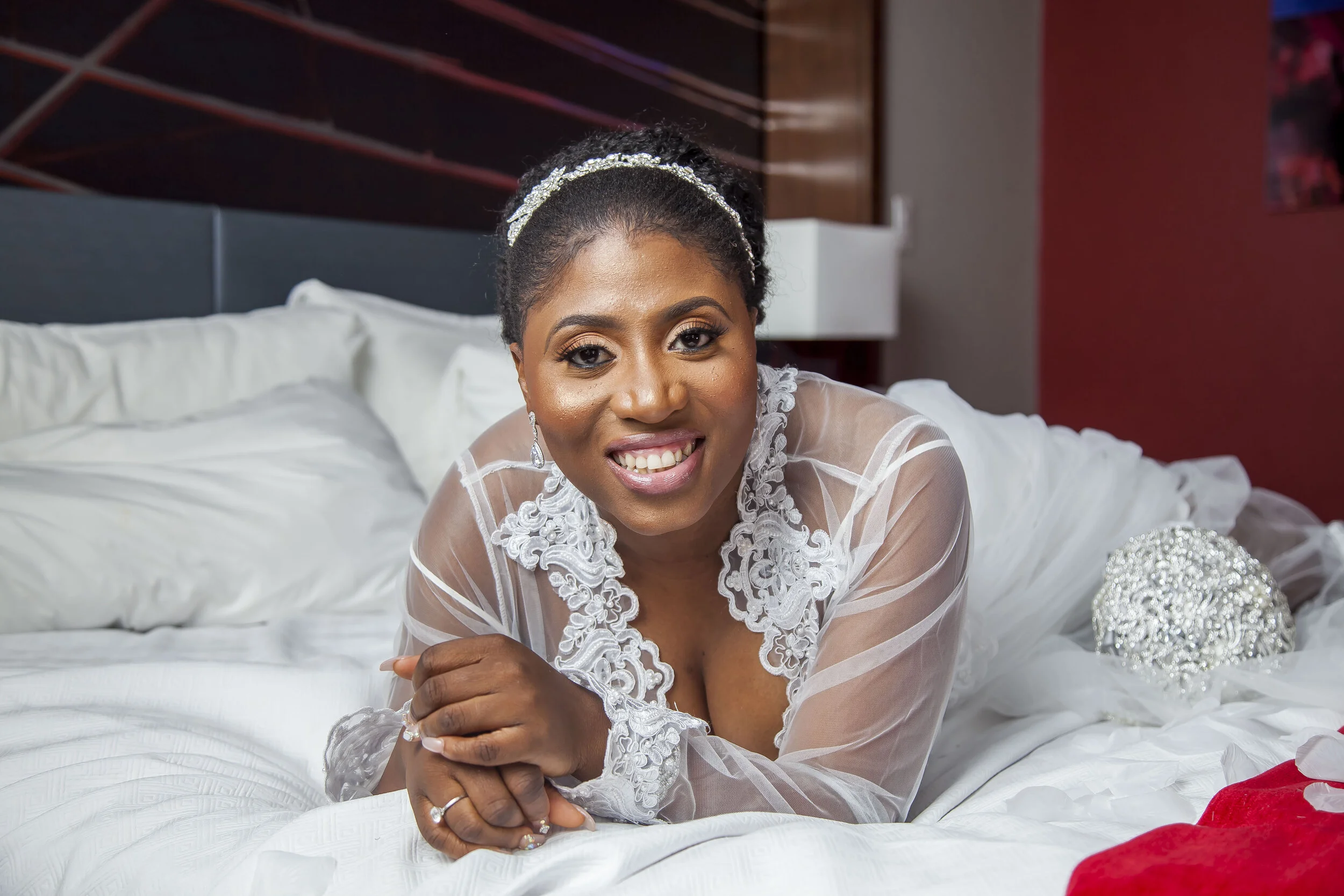A smiling woman in a wedding dress lying on a bed with white bedding, holding her hands together, with a decorative headband, earrings, and makeup, in a modern room with dark and red walls.