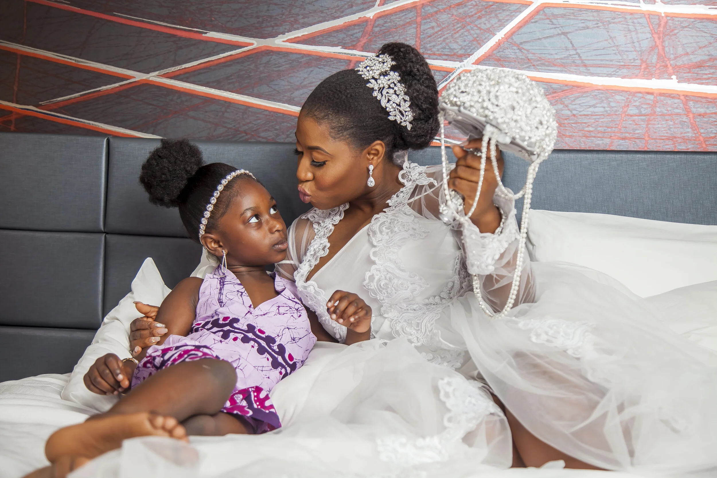 A woman in a white wedding dress with lace details gently holding a young girl in purple and black pajamas, both with elegant hairstyles adorned with sparkling hair accessories, sitting on a bed with a dark padded headboard against a colorful abstrac