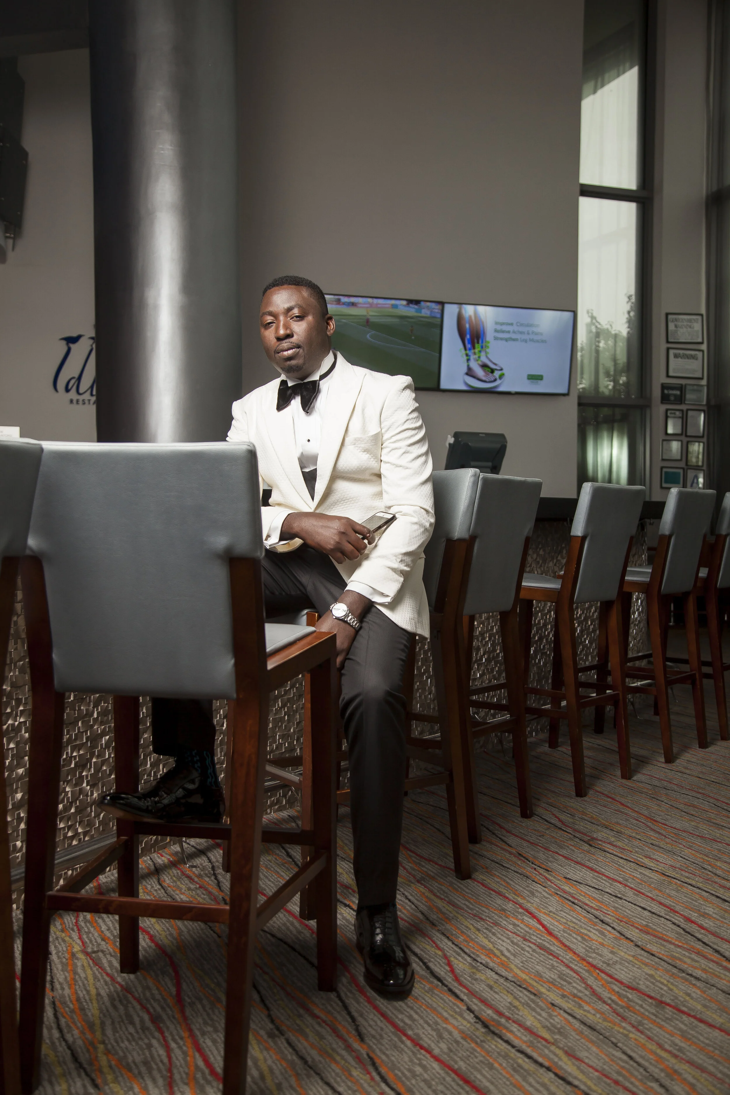 A man in a white tuxedo with a black bow tie sitting at a table in a restaurant or conference room, with a large television screen and windows in the background.