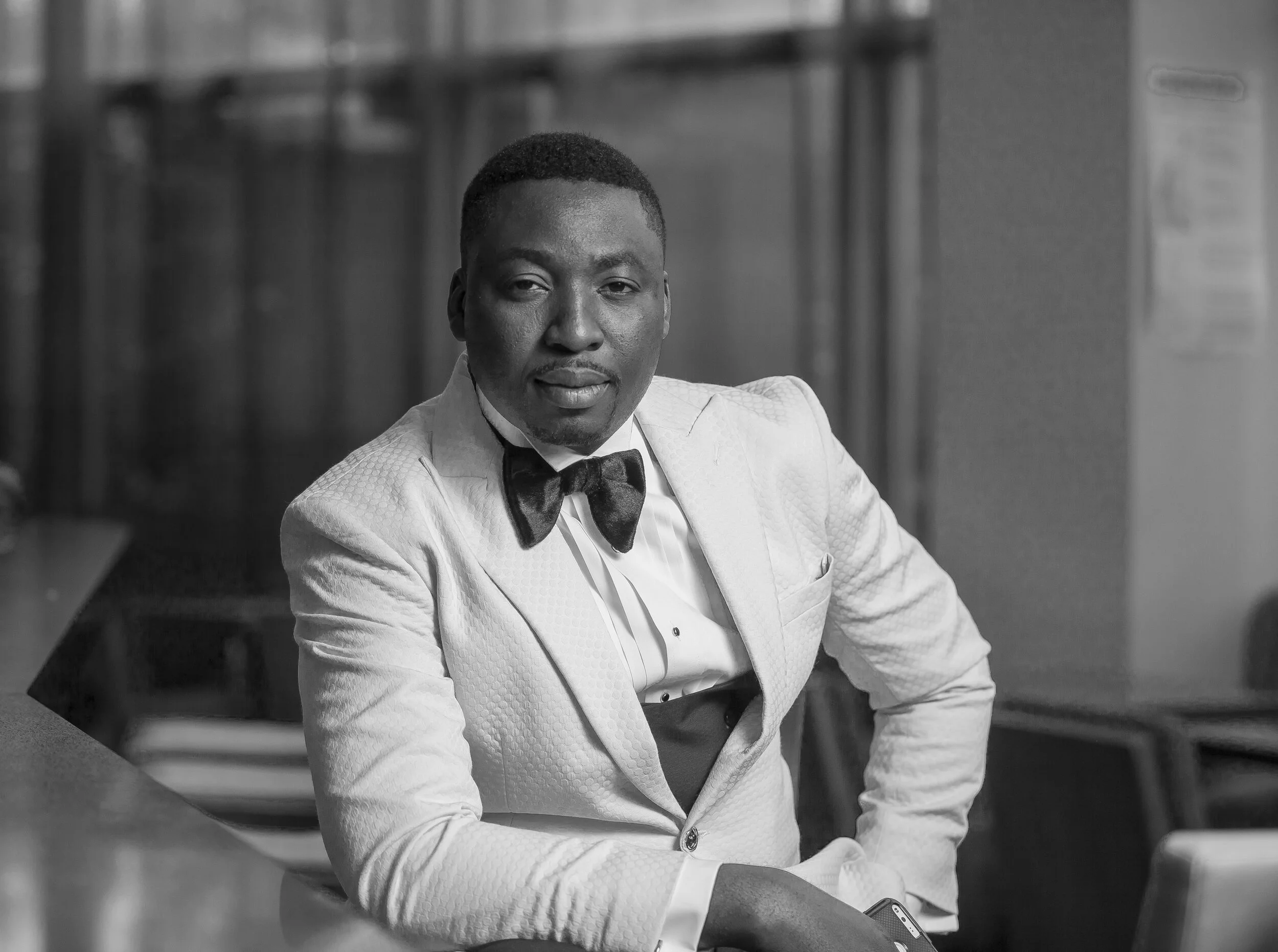 Man in a white tuxedo with a black bow tie sitting at a table, looking at the camera, in a formal setting