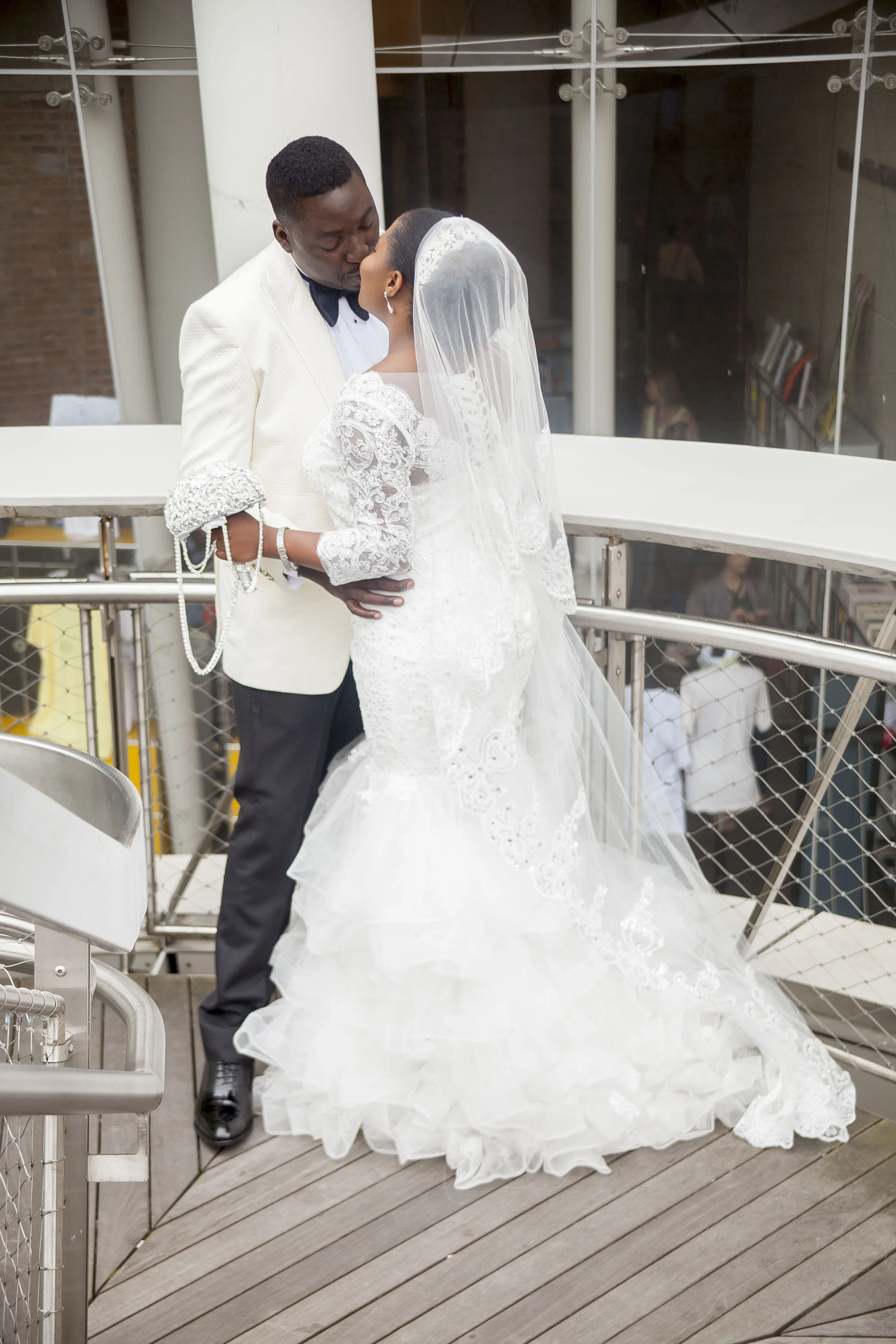 A bride and groom share a kiss during their wedding ceremony on an outdoor terrace, with the bride wearing a white lace wedding dress and veil, and the groom dressed in a white tuxedo jacket with a black bow tie.