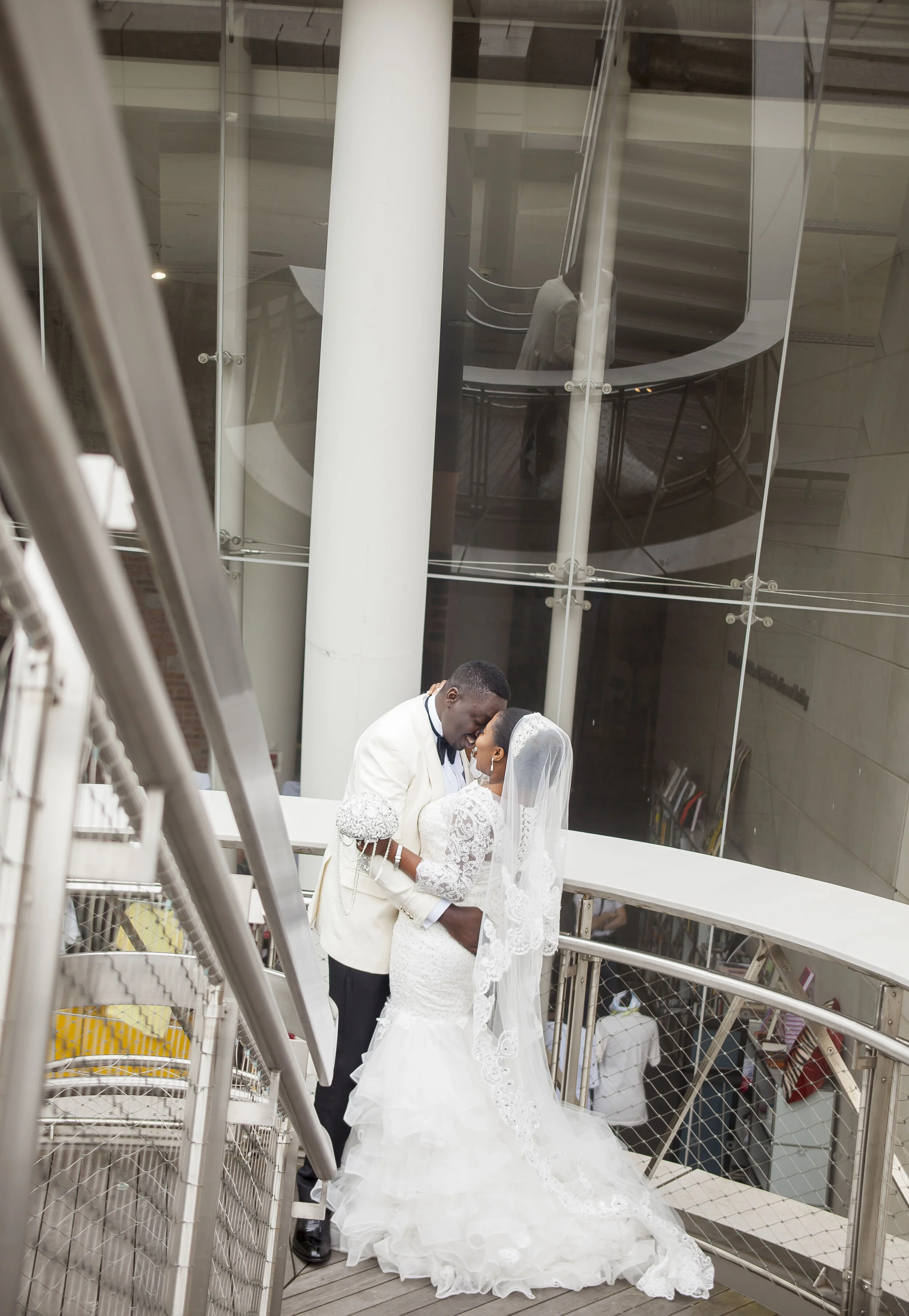 A bride and groom share a kiss on an outdoor walkway with a glass building and modern staircase reflected in the background.