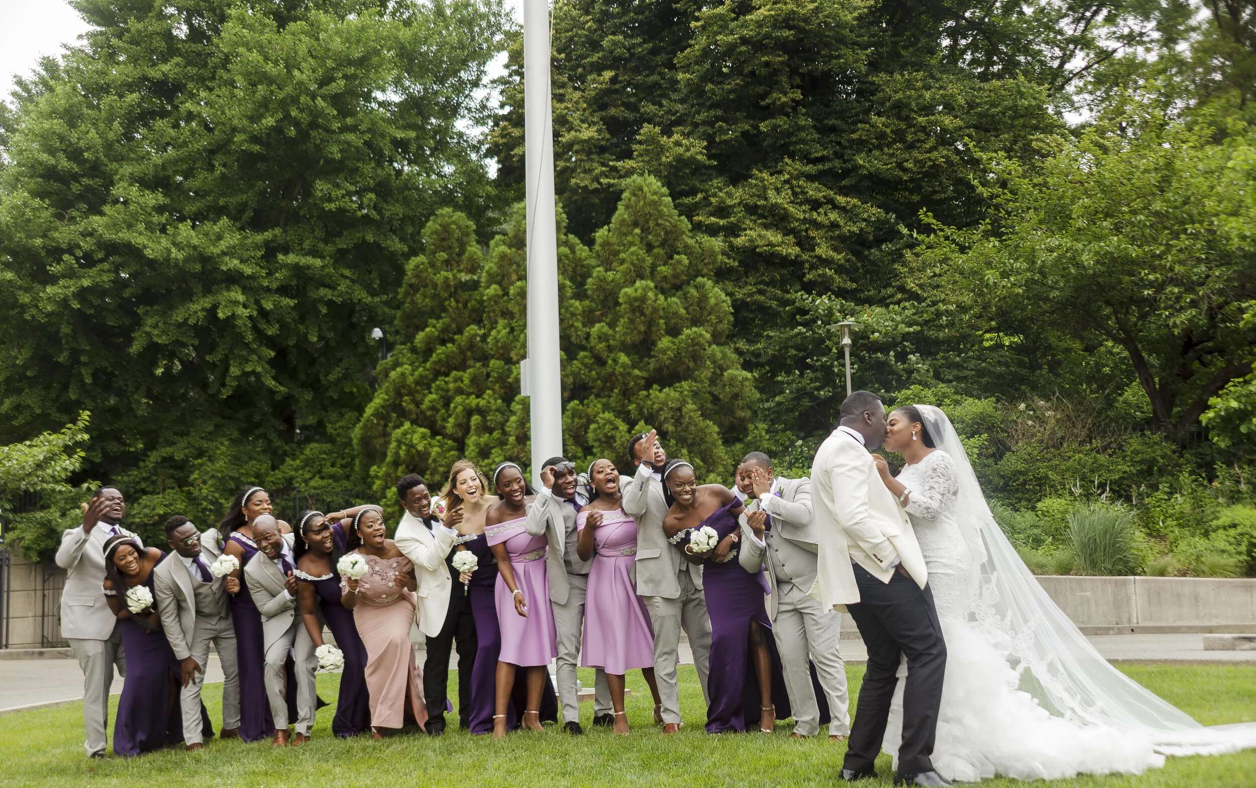 A group of wedding guests and the bride and groom outdoors, surrounded by green trees, celebrating the wedding with happy expressions and laughter.