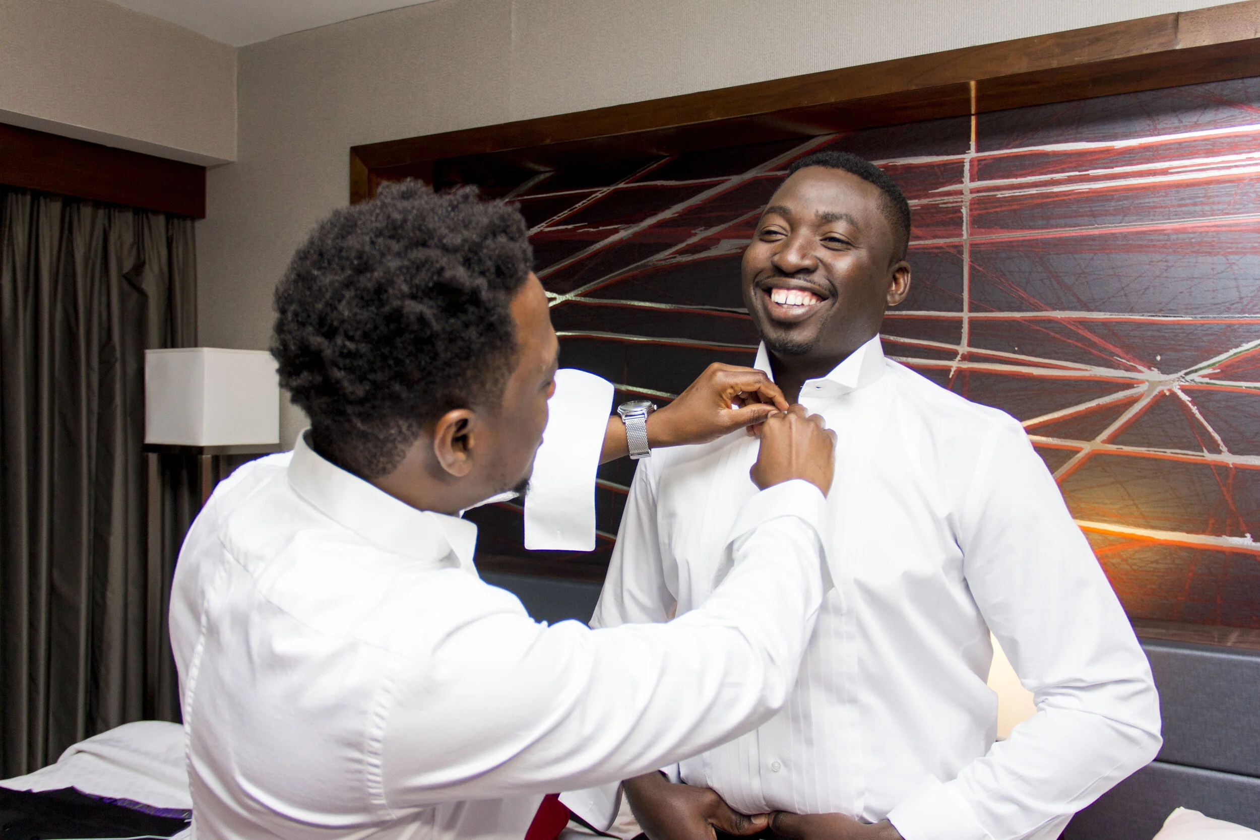 Two men in white dress shirts, one helping the other with a tie, in a hotel room with abstract wall art.