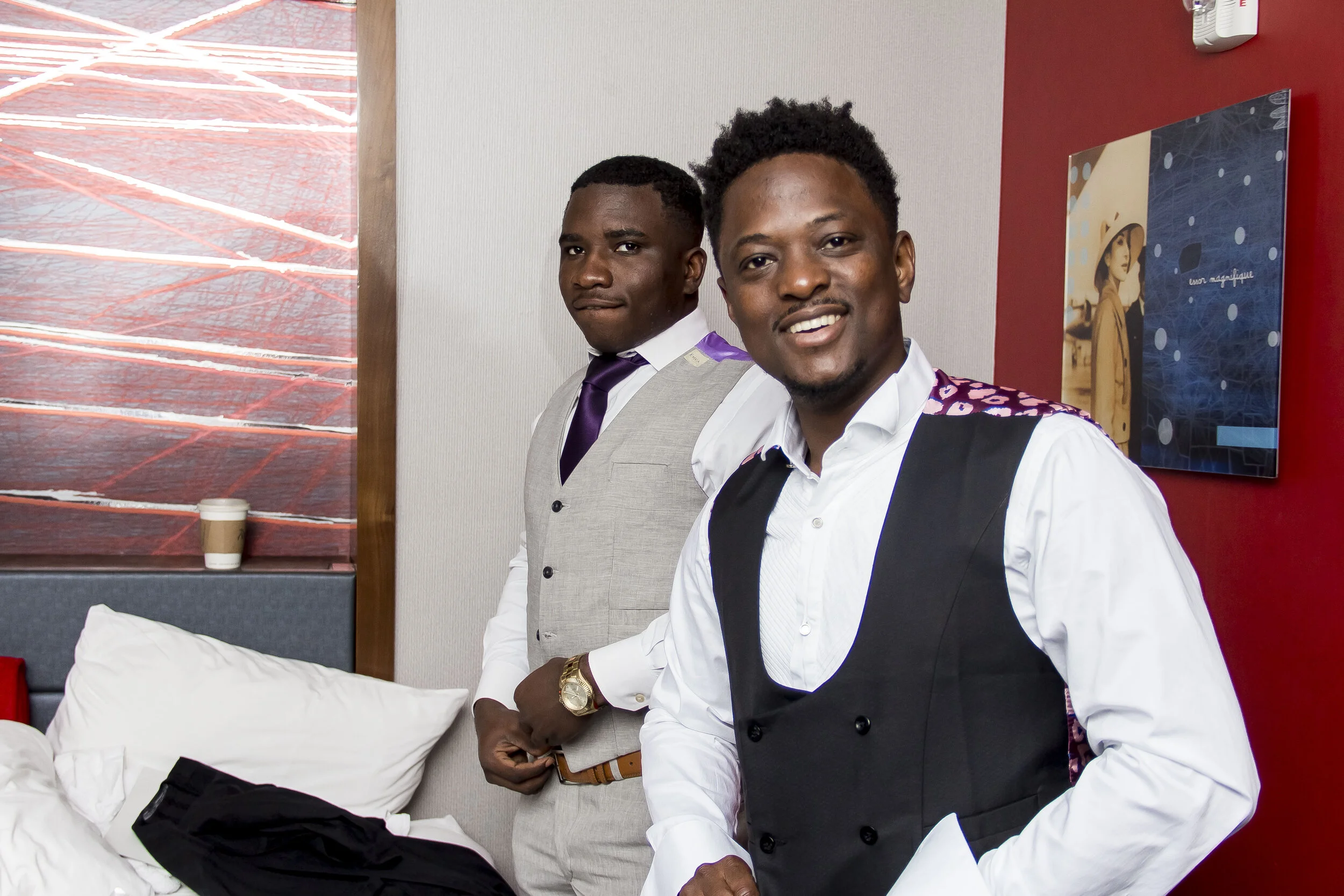 Two young men dressed in formal attire standing indoors, one wearing a light gray suit with a purple tie and the other wearing a white shirt with a black vest and colorful patterned jacket, smiling at the camera. There are posters on the wall and a b