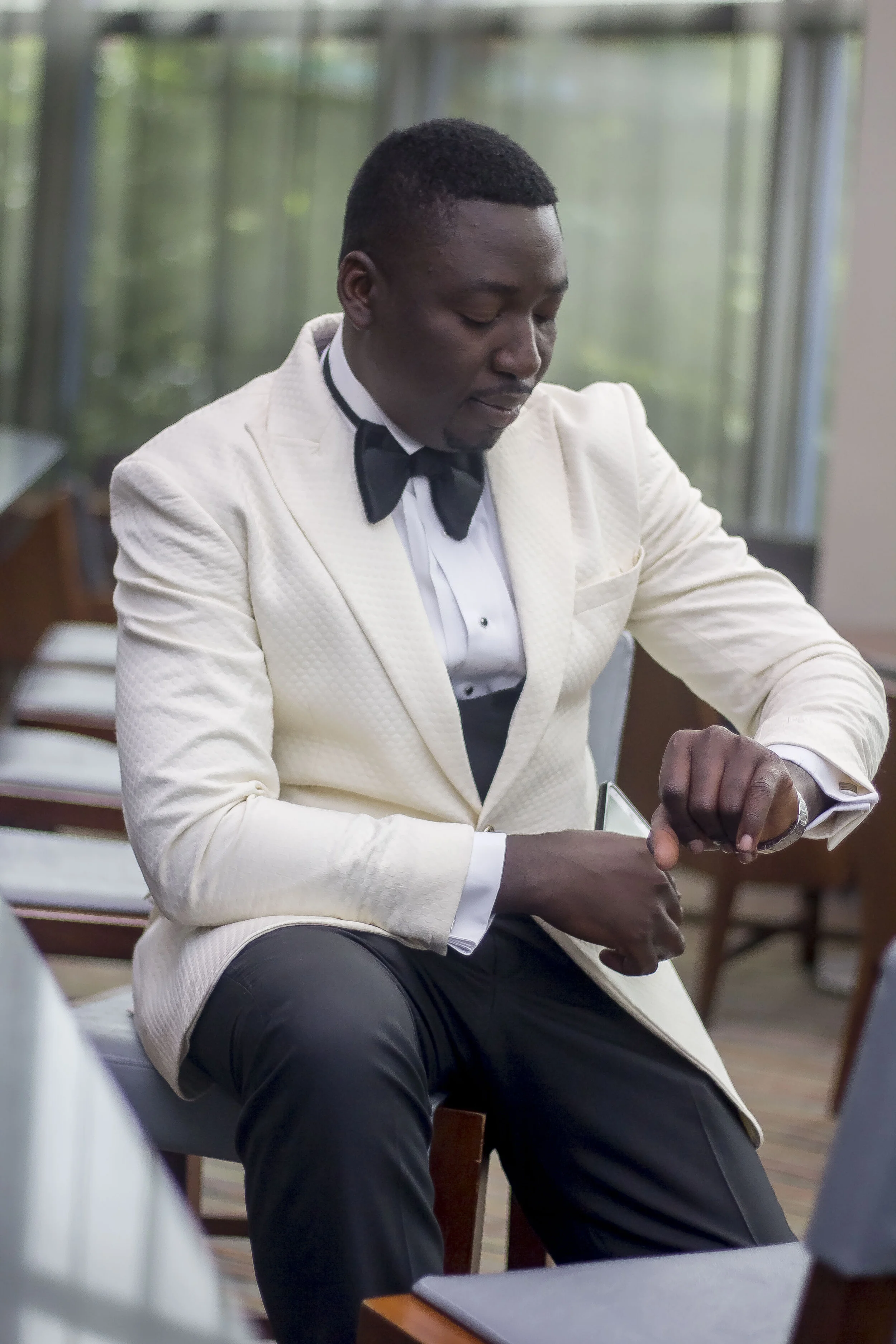 A handsome young man with dark skin and short hair is wearing a white tuxedo with a black bow tie. He is sitting in a room with large windows and is looking at his watch.