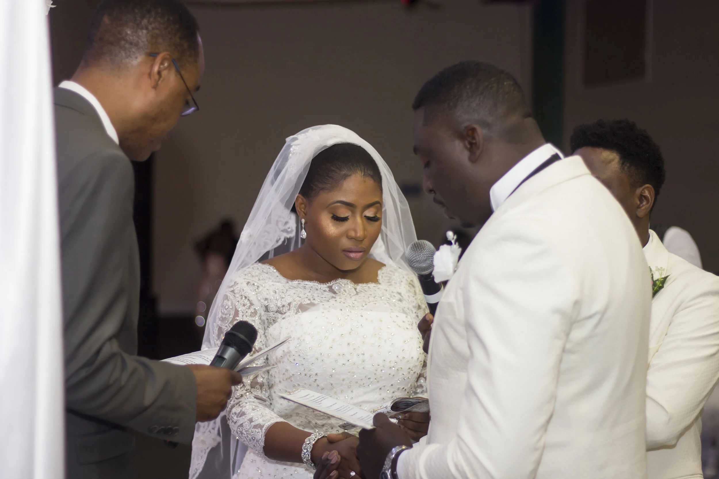 A bride and groom standing face to face during their wedding ceremony, holding hands and with eyes closed, surrounded by officiant and witnesses.
