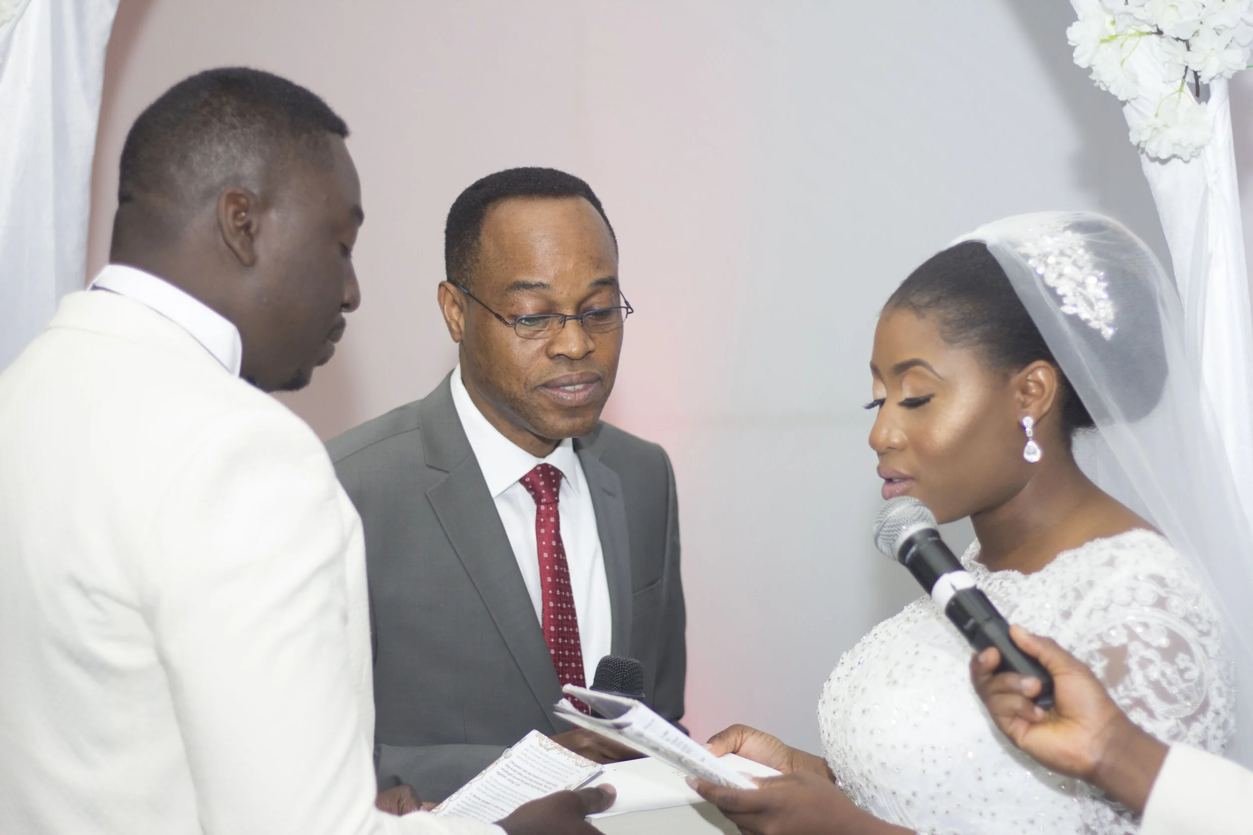 A wedding ceremony with a bride and groom exchanging rings, with an officiant and a person holding a microphone.