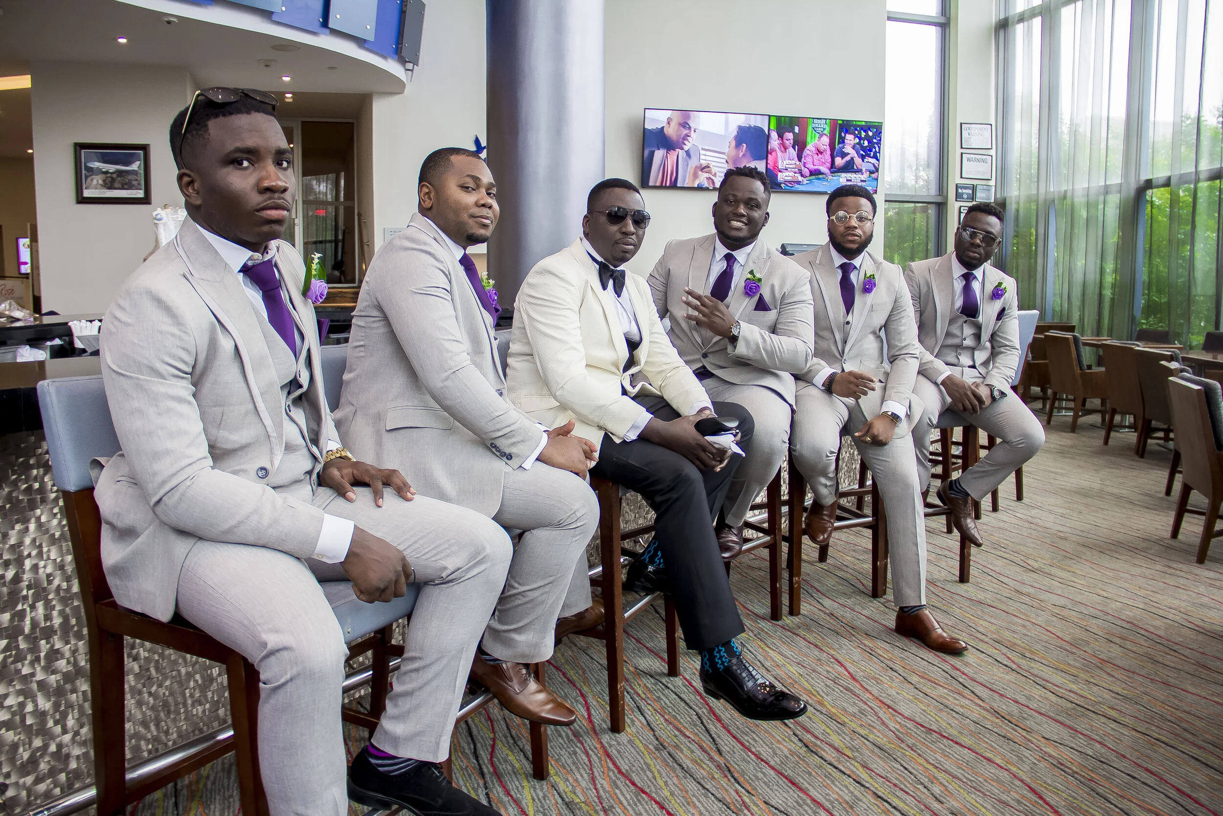 A group of seven men dressed in light-colored suits and purple ties, some with purple flowers pinning on their lapels, sitting on stools in a bright indoor space with large windows. They are posing for the photo, with some looking directly at the cam